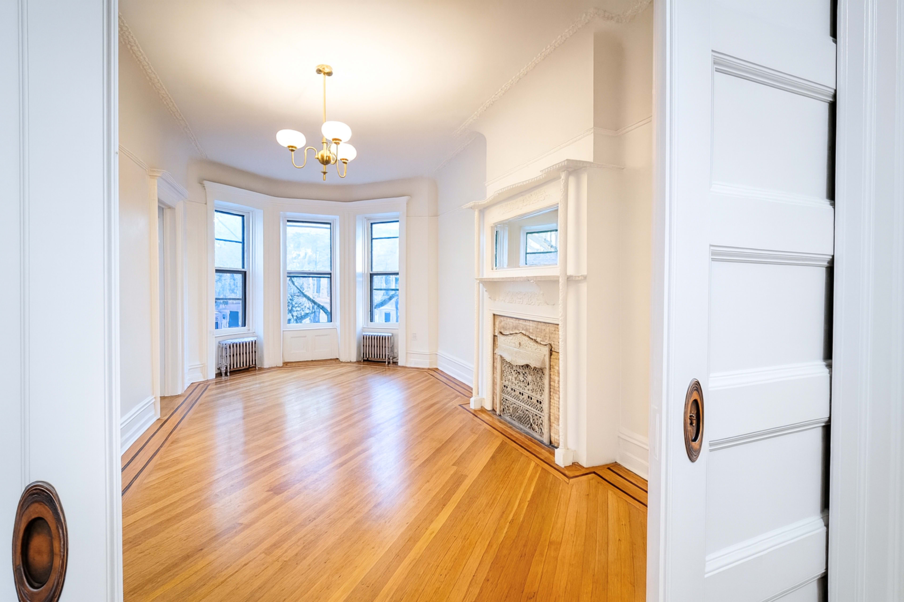 The image shows a bright, empty room with large windows, hardwood floors, and a decorative fireplace.