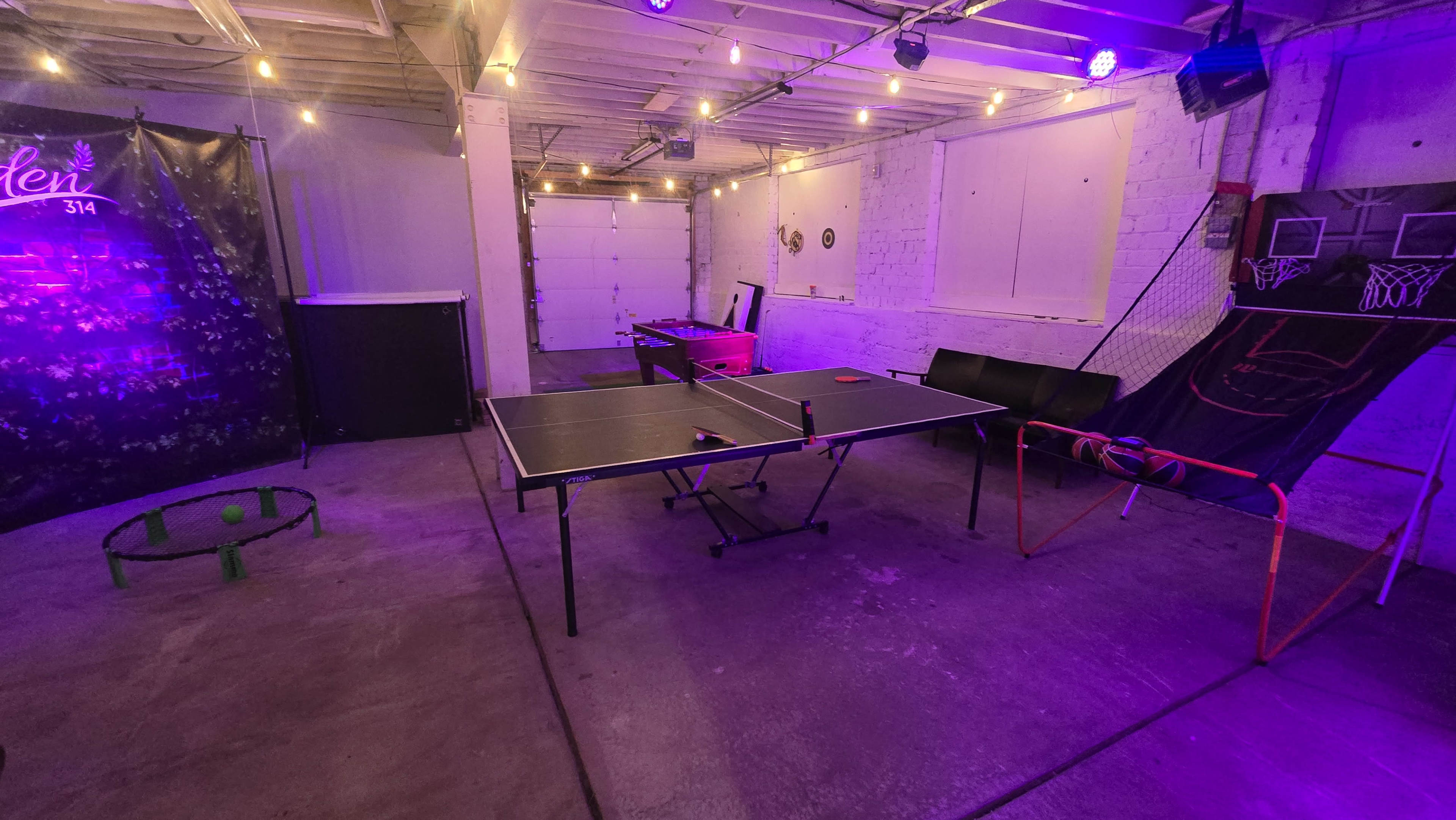 The image shows a recreational room featuring a ping pong table, a foosball table, and sports-themed decor under dim lighting with string lights.