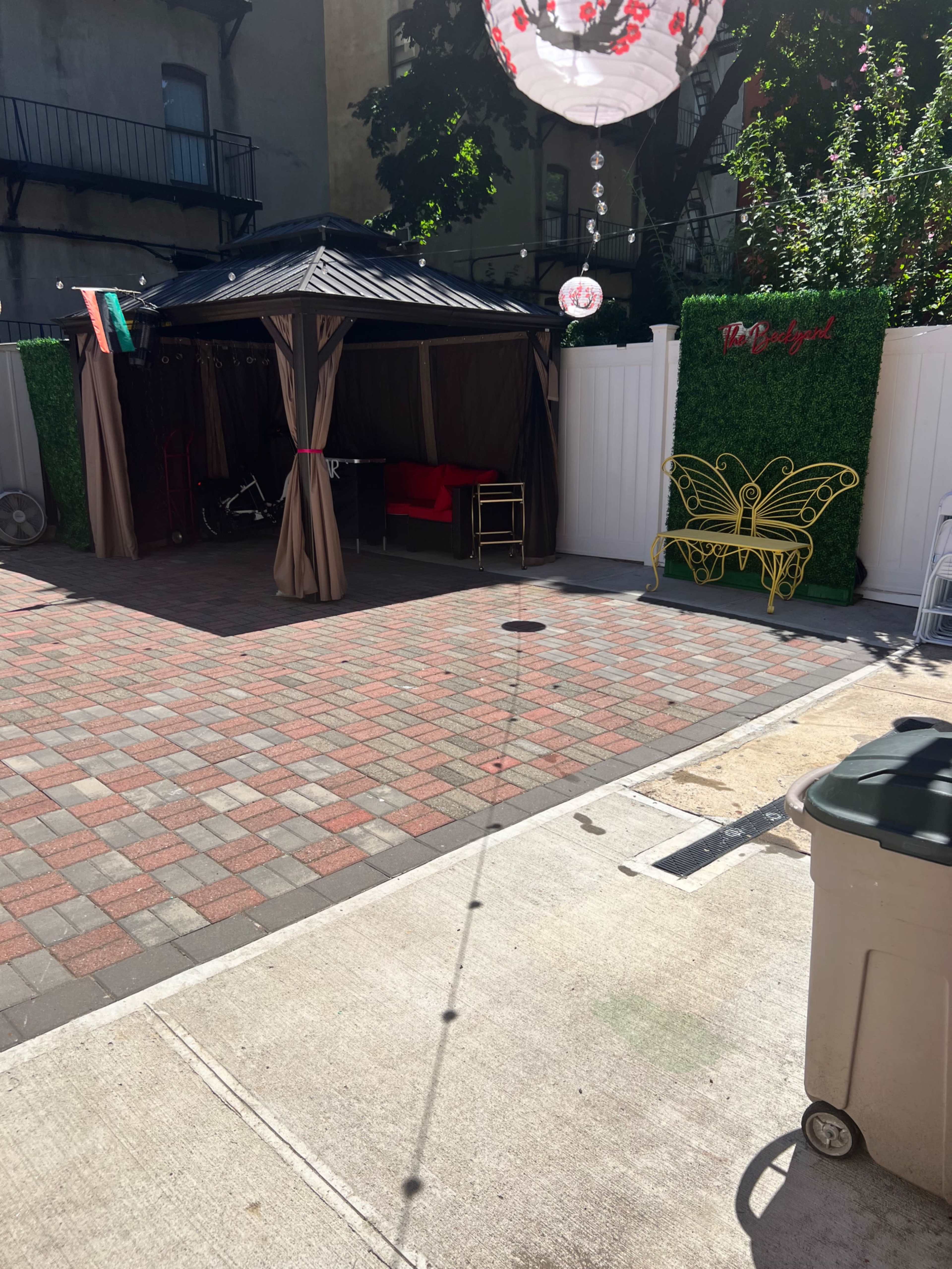A small outdoor patio area with a gazebo, a red couch, a green decorative wall, and a patterned brick pathway.