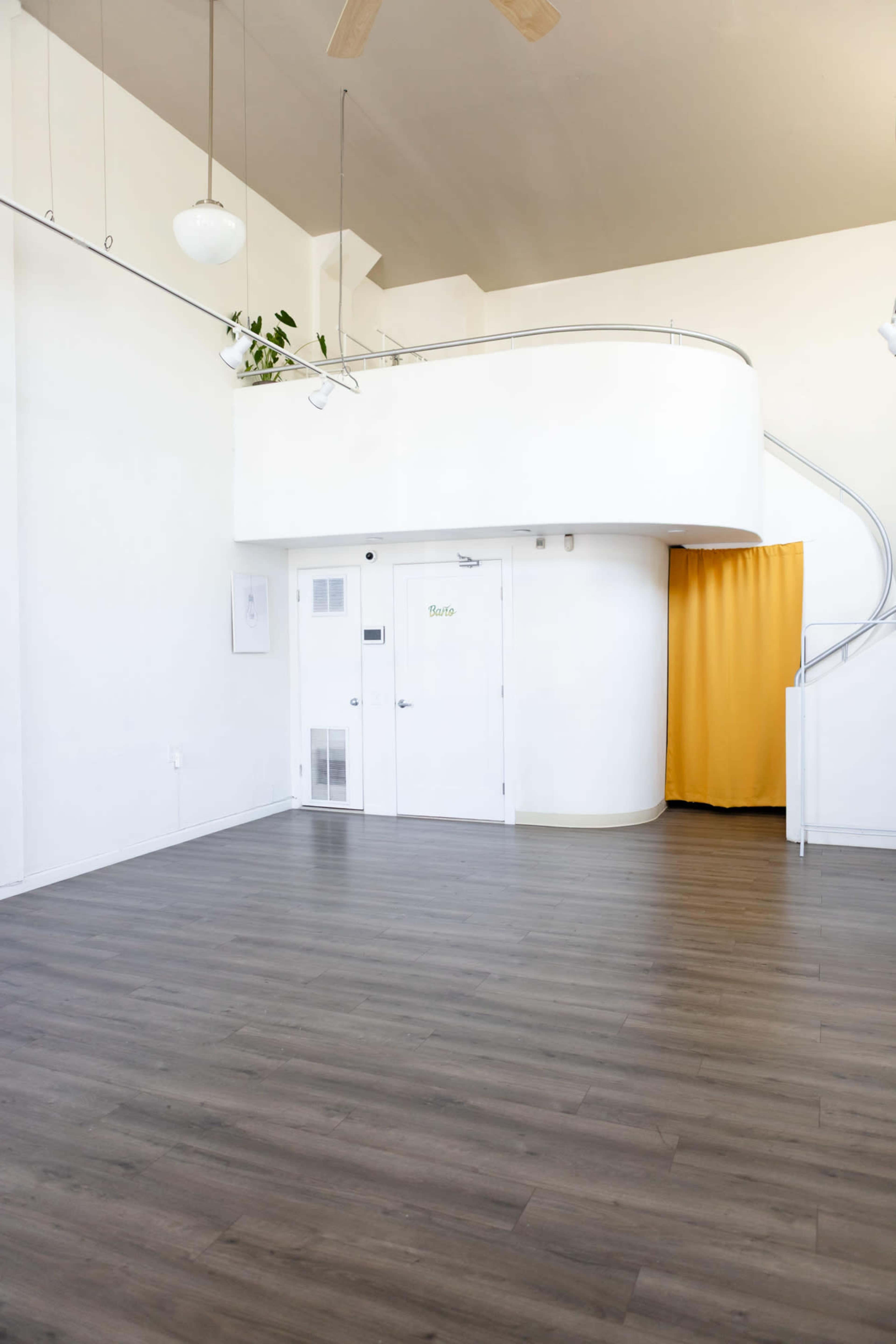 The image shows a spacious interior with a wooden floor, a white wall featuring a door labeled "Studio," and a curved staircase leading to an upper level, complemented by a yellow curtain.