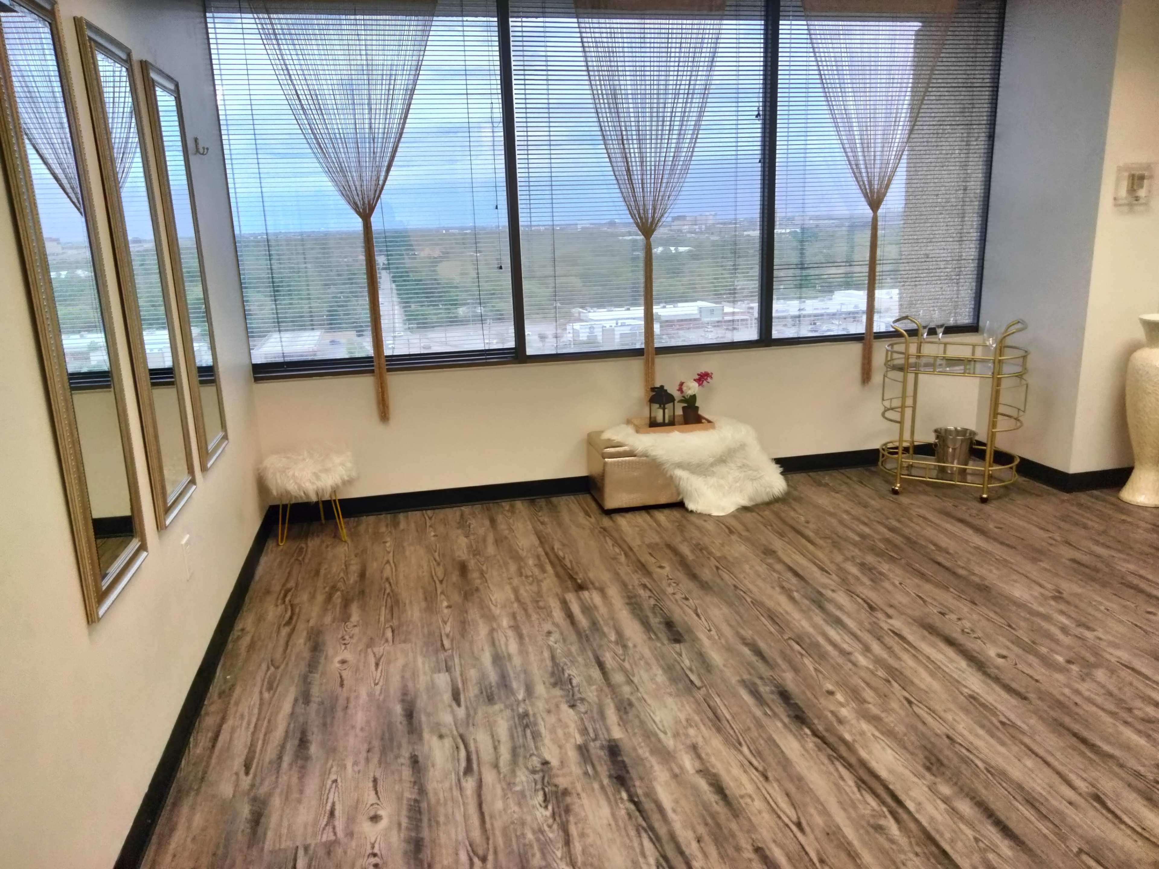 An interior space with wooden floors, large windows covered by light curtains, three mirrors, a small white stool, a decorative cart, and a white faux fur throw on a box.