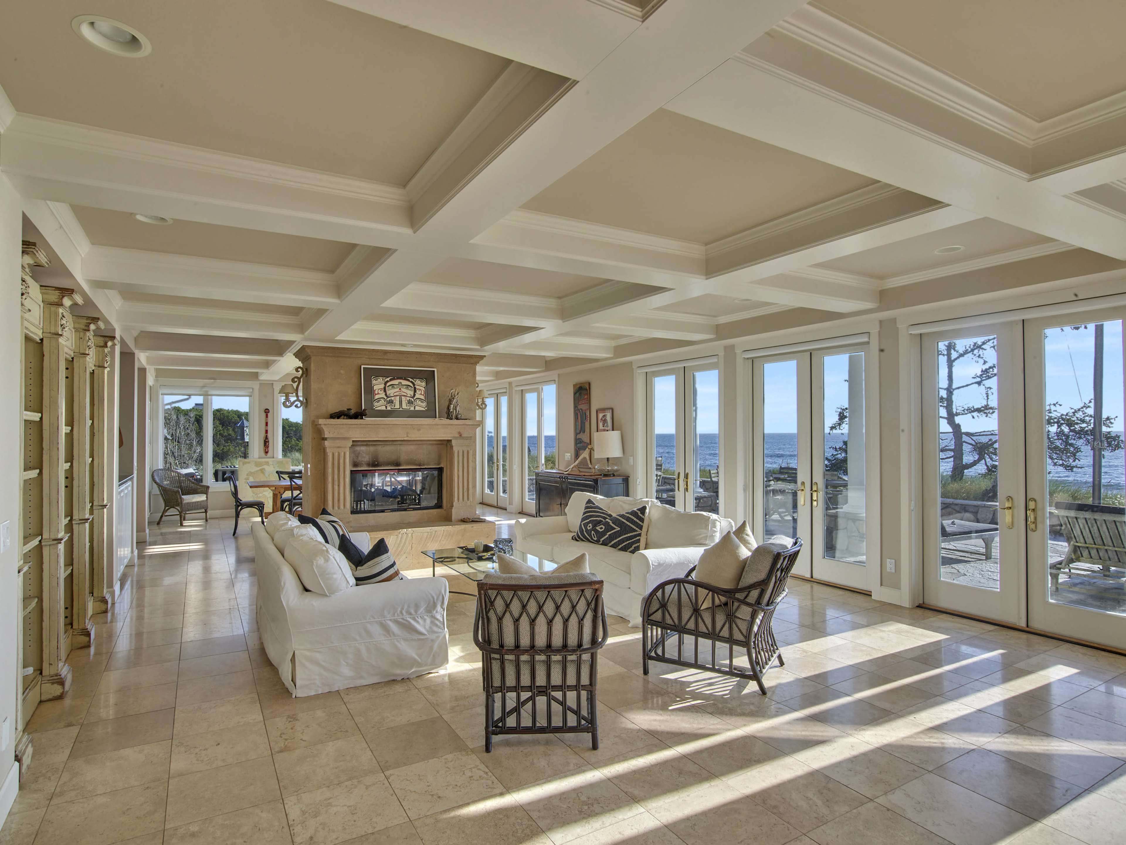 The spacious living room features large windows with a view of the ocean, a stone fireplace, and light-colored furniture arranged for comfort.