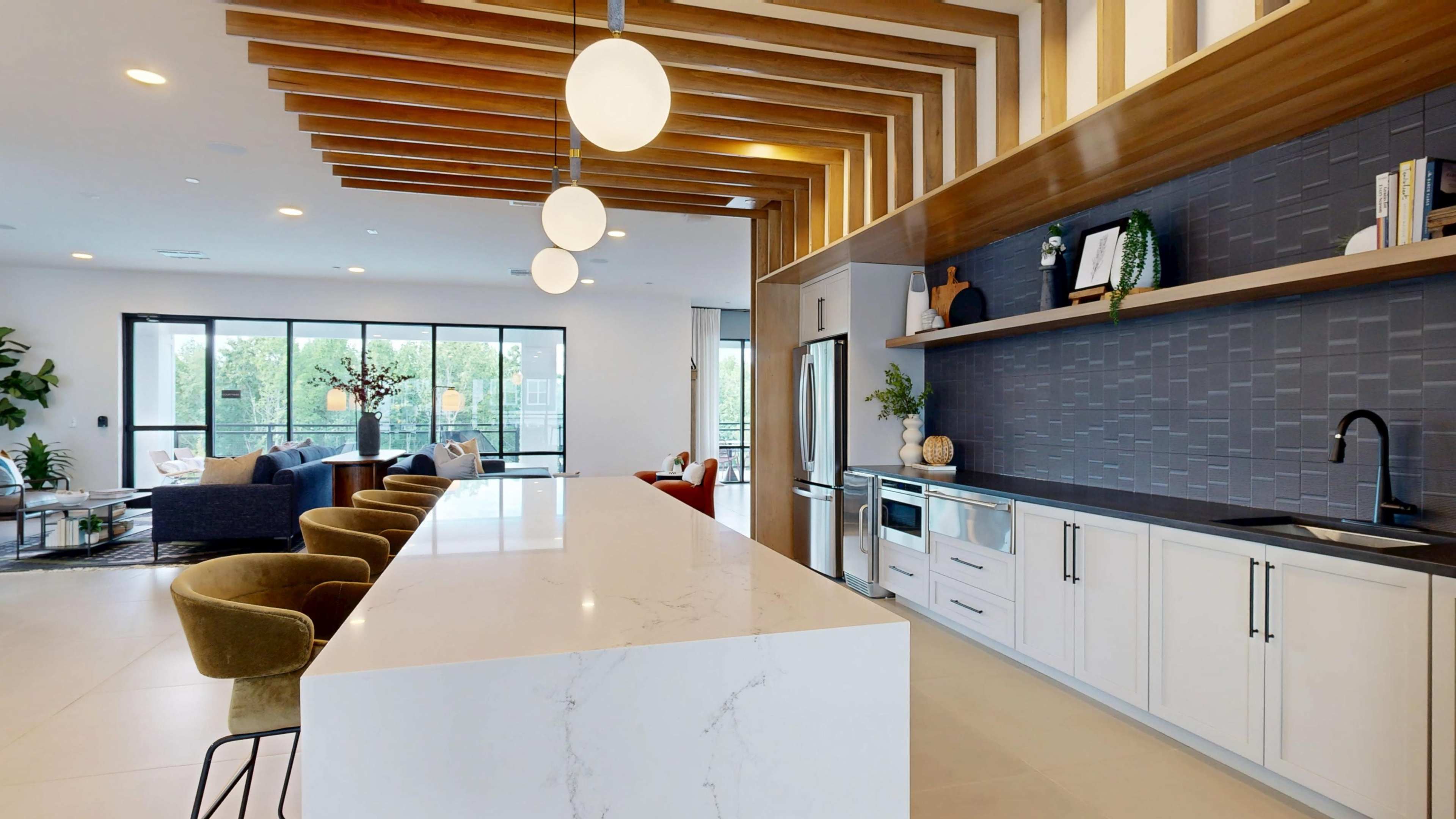 The image shows a modern kitchen and living area featuring a large white island, wooden ceiling beams, and sleek cabinetry with a shelf displaying decorative items.