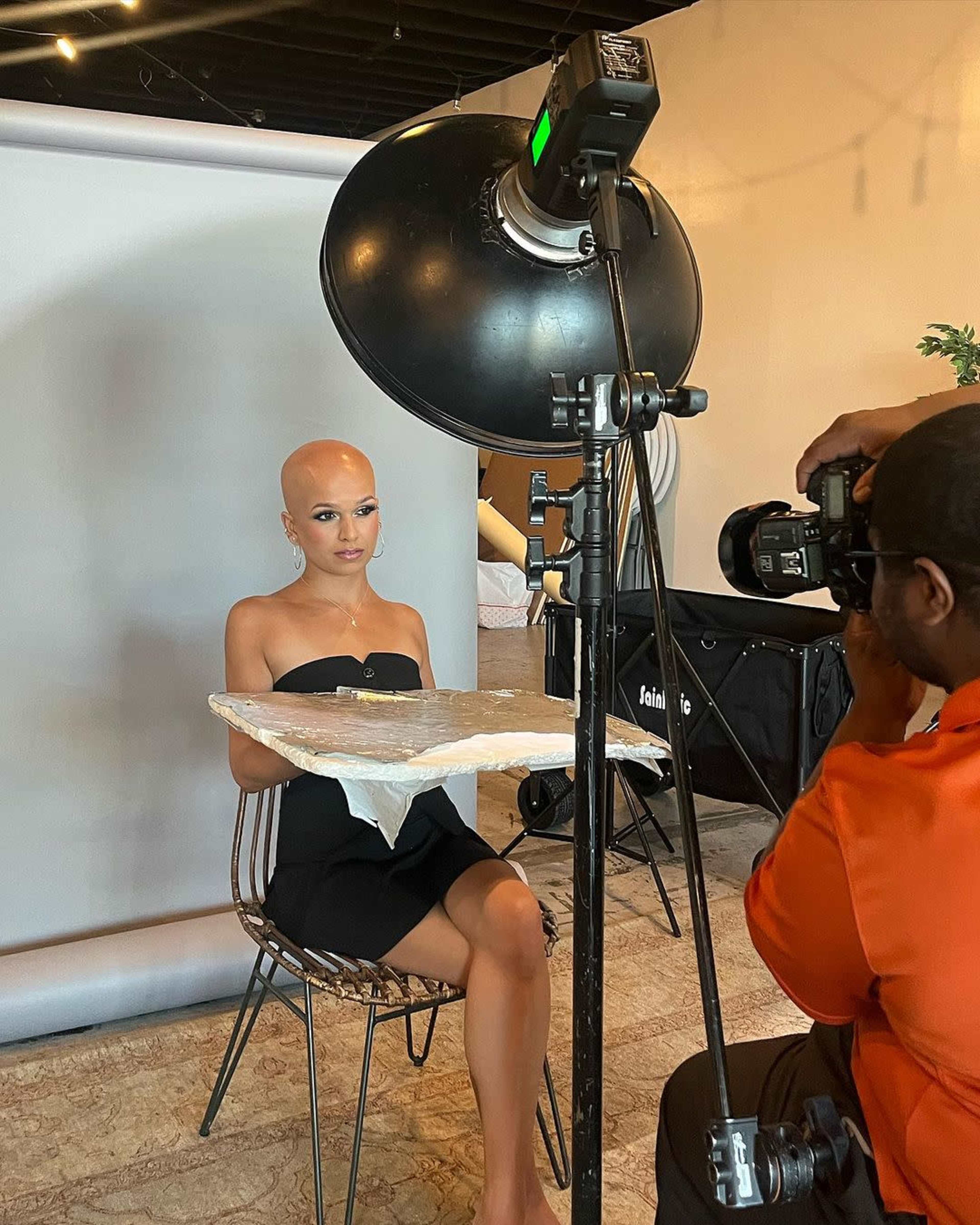 A bald model sits on a chair holding a plate while a photographer captures her image in a studio setting.