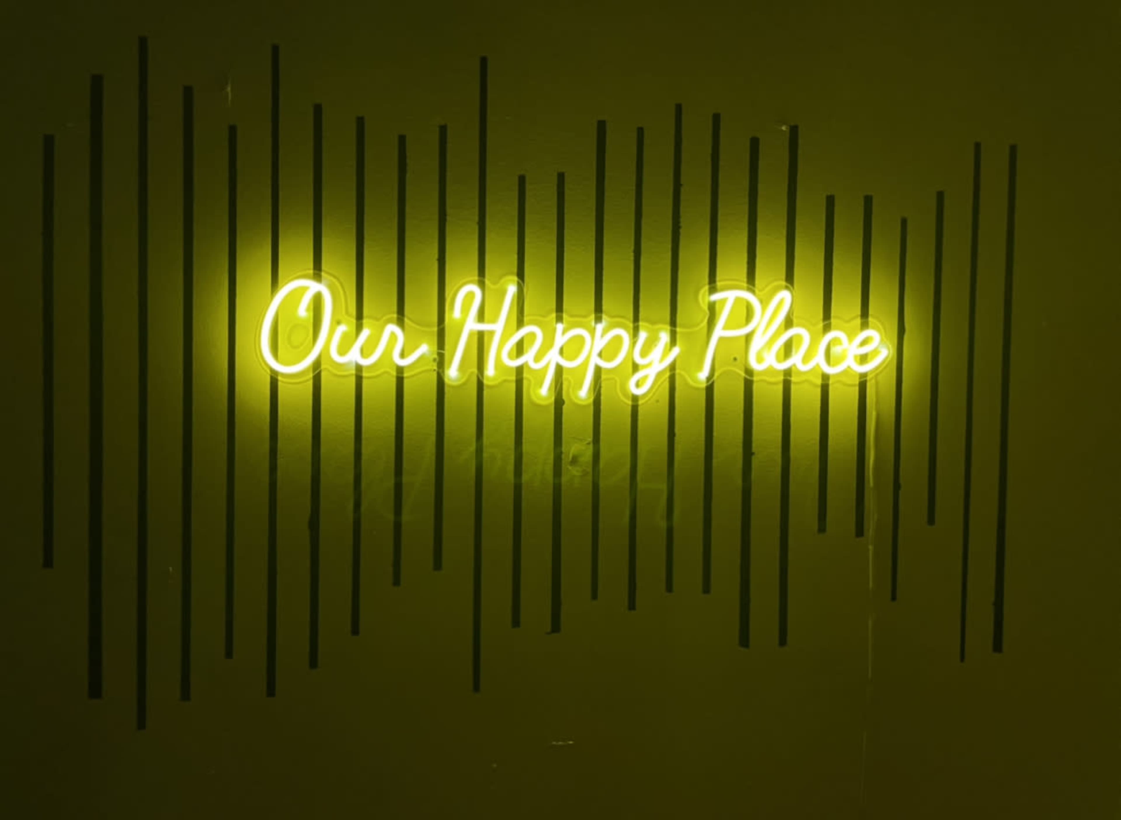 A neon sign that reads "Our Happy Place" is illuminated against a dark green wall with linear designs.