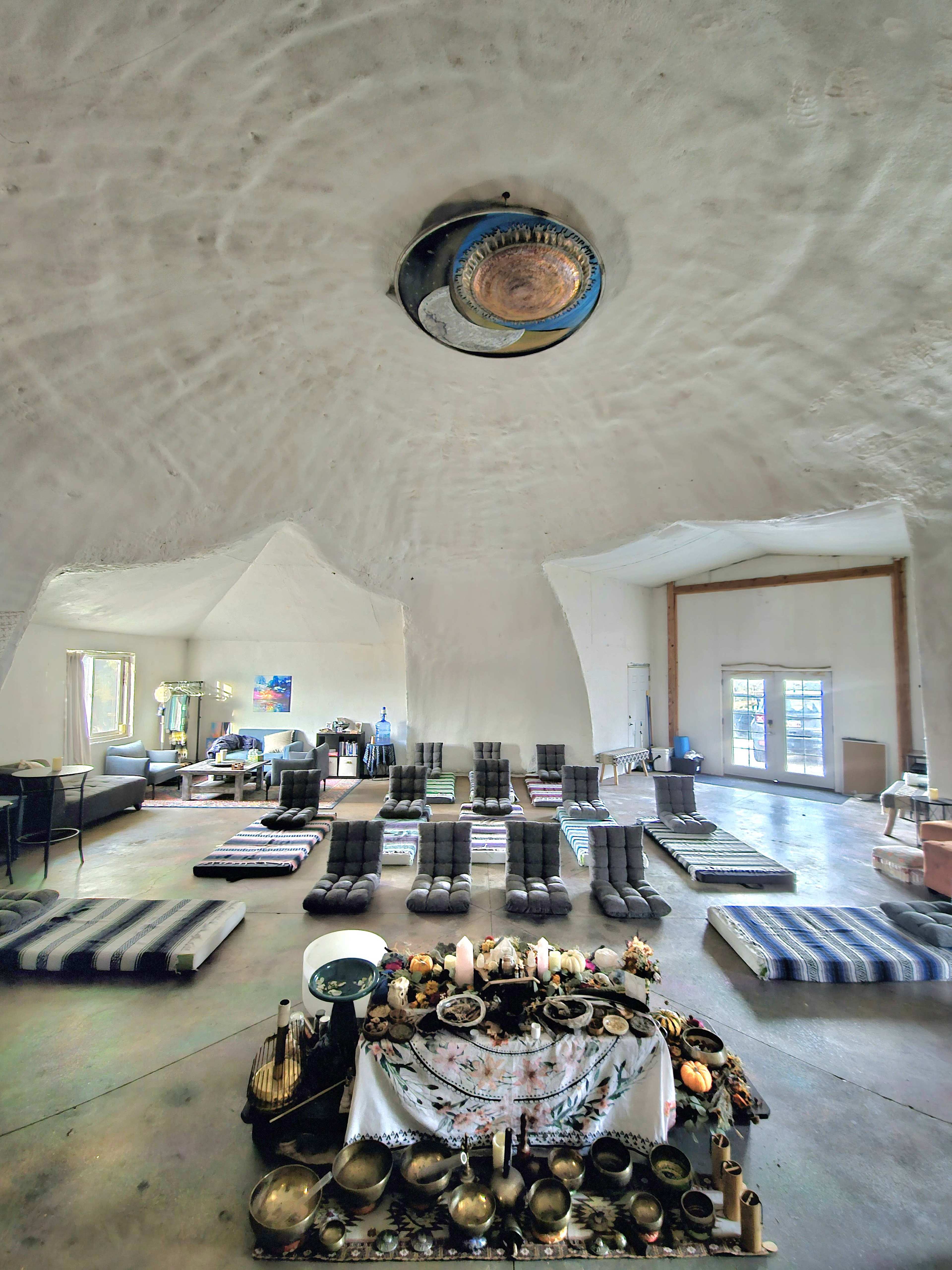 The image shows a spacious, dome-shaped interior with arranged meditation mats and an altar featuring various objects at the center.