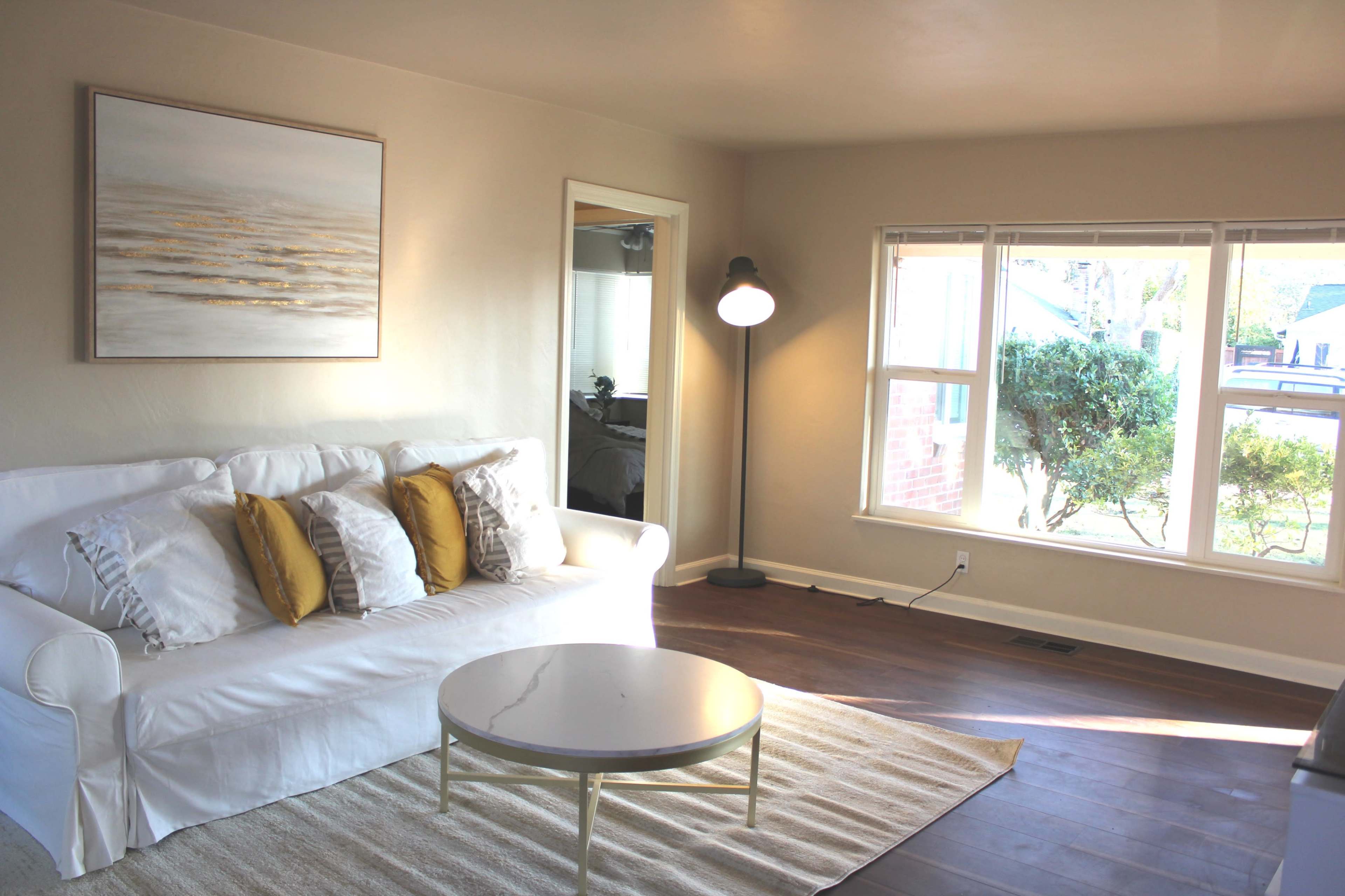 A bright living room features a white sofa with yellow accent pillows, a round coffee table, a floor lamp, and large windows allowing natural light to illuminate the space.
