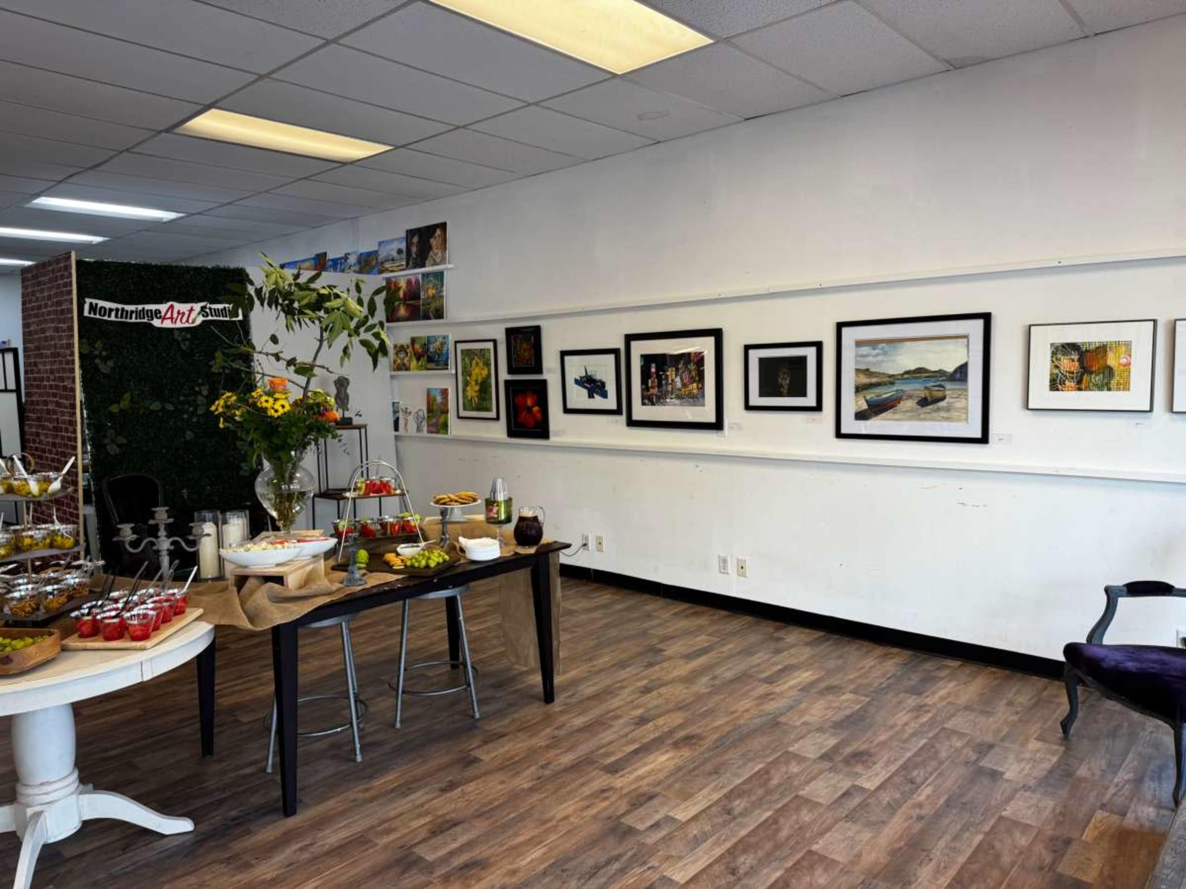 The image shows an art gallery setting with framed artworks hanging on the walls and a table displaying food and beverages for an event.