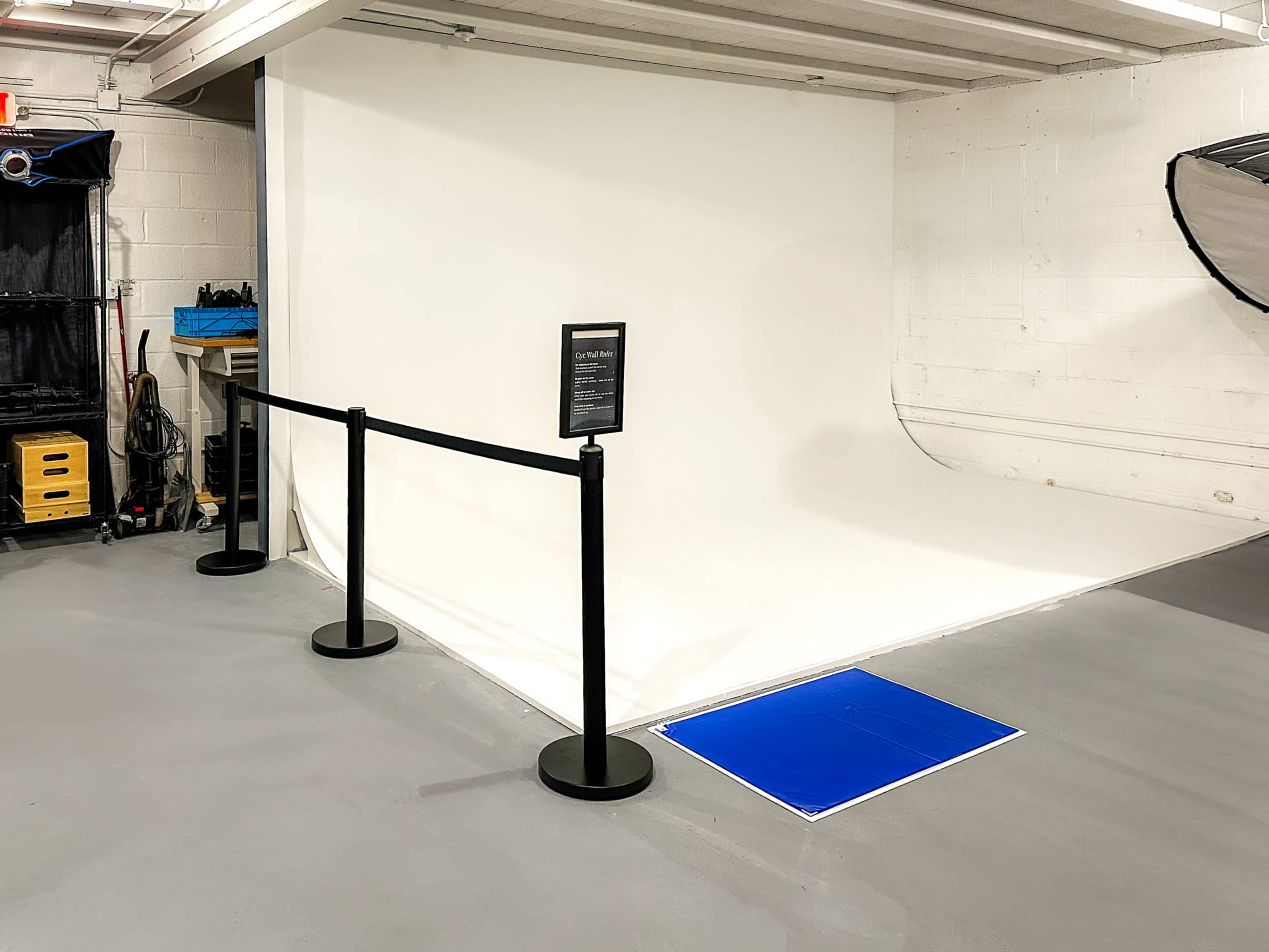 The image shows a photography studio with a white backdrop, a blue mat on the floor, and a rope barrier in front of it.