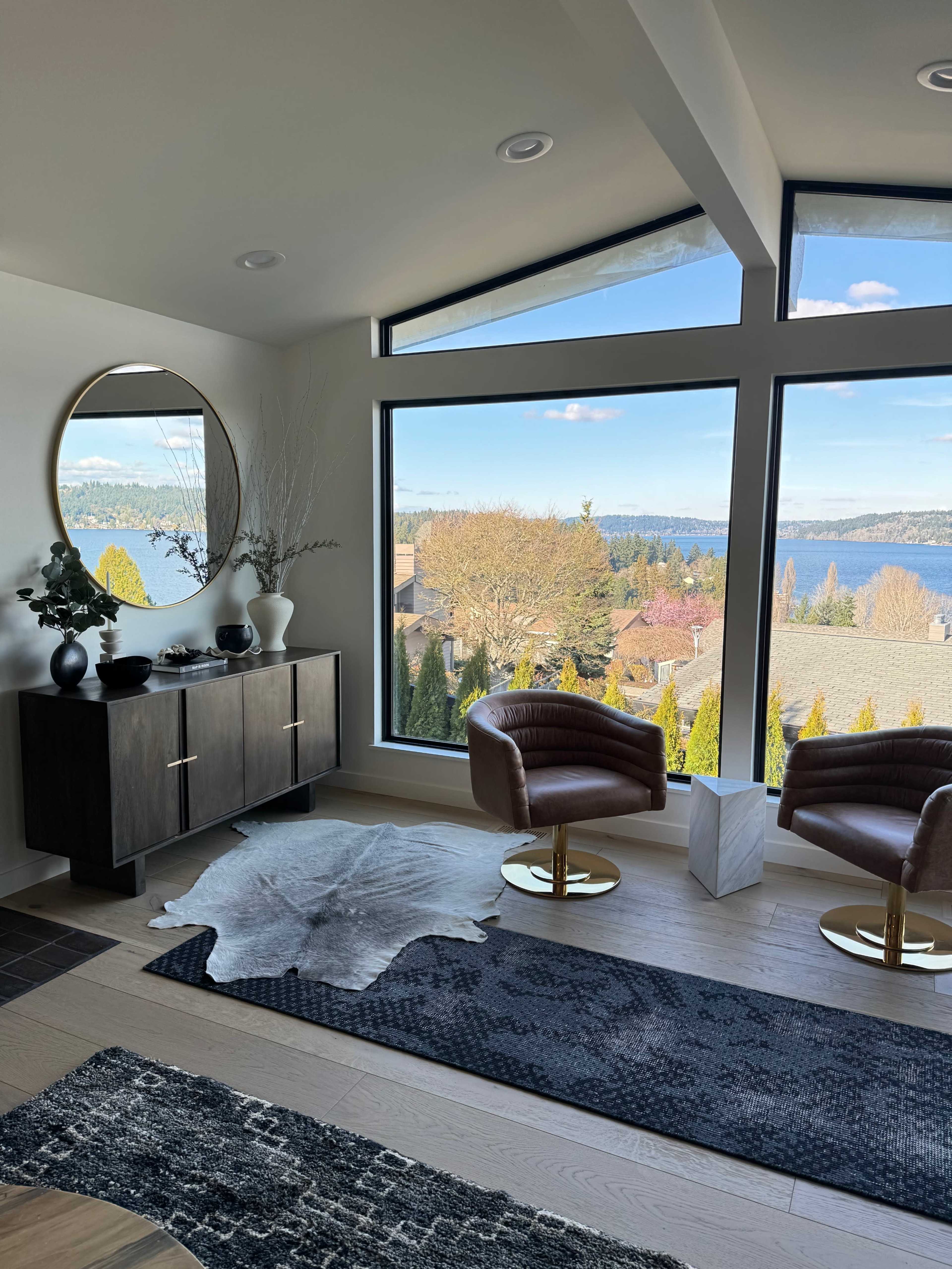 The image shows a modern interior space with two brown chairs, a dark wooden sideboard, and a large window offering a view of the outside landscape and water.