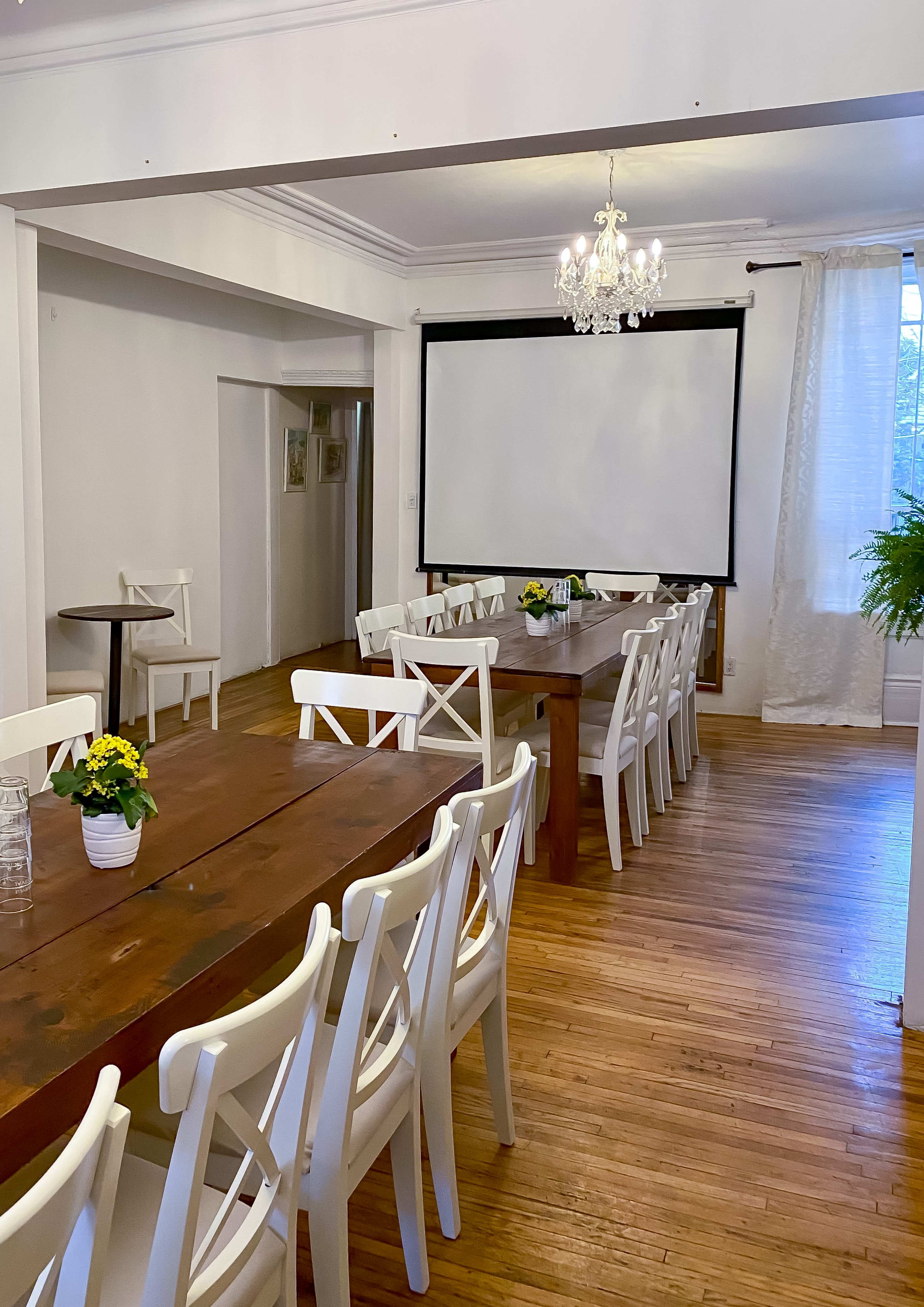 A meeting room features two long wooden tables surrounded by white chairs, with a large screen at one end and a chandelier overhead.