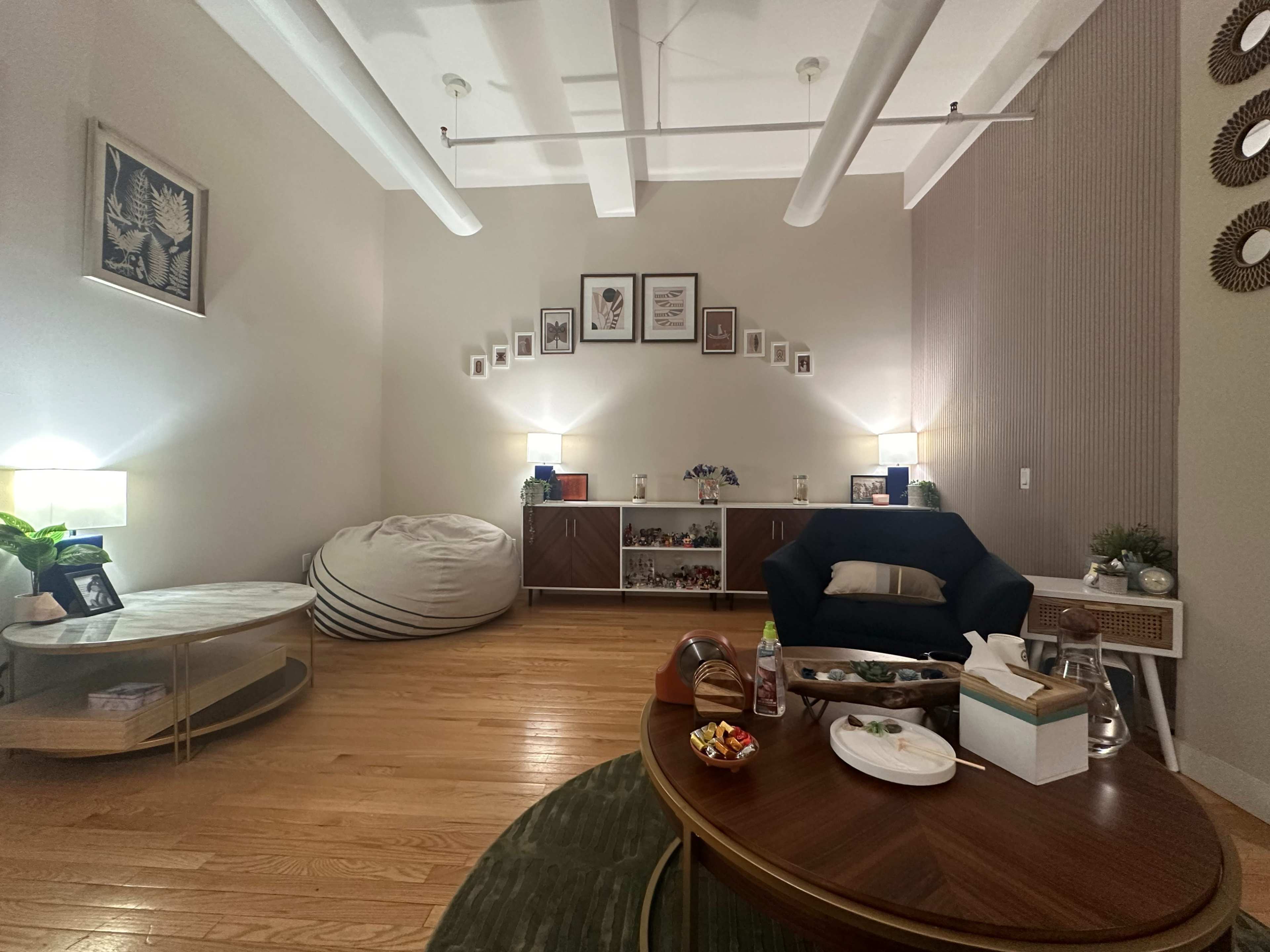 The image shows a cozy room with wooden flooring, featuring a large bean bag, a dark armchair, a circular coffee table with snacks, and shelves adorned with framed pictures and decorative items.