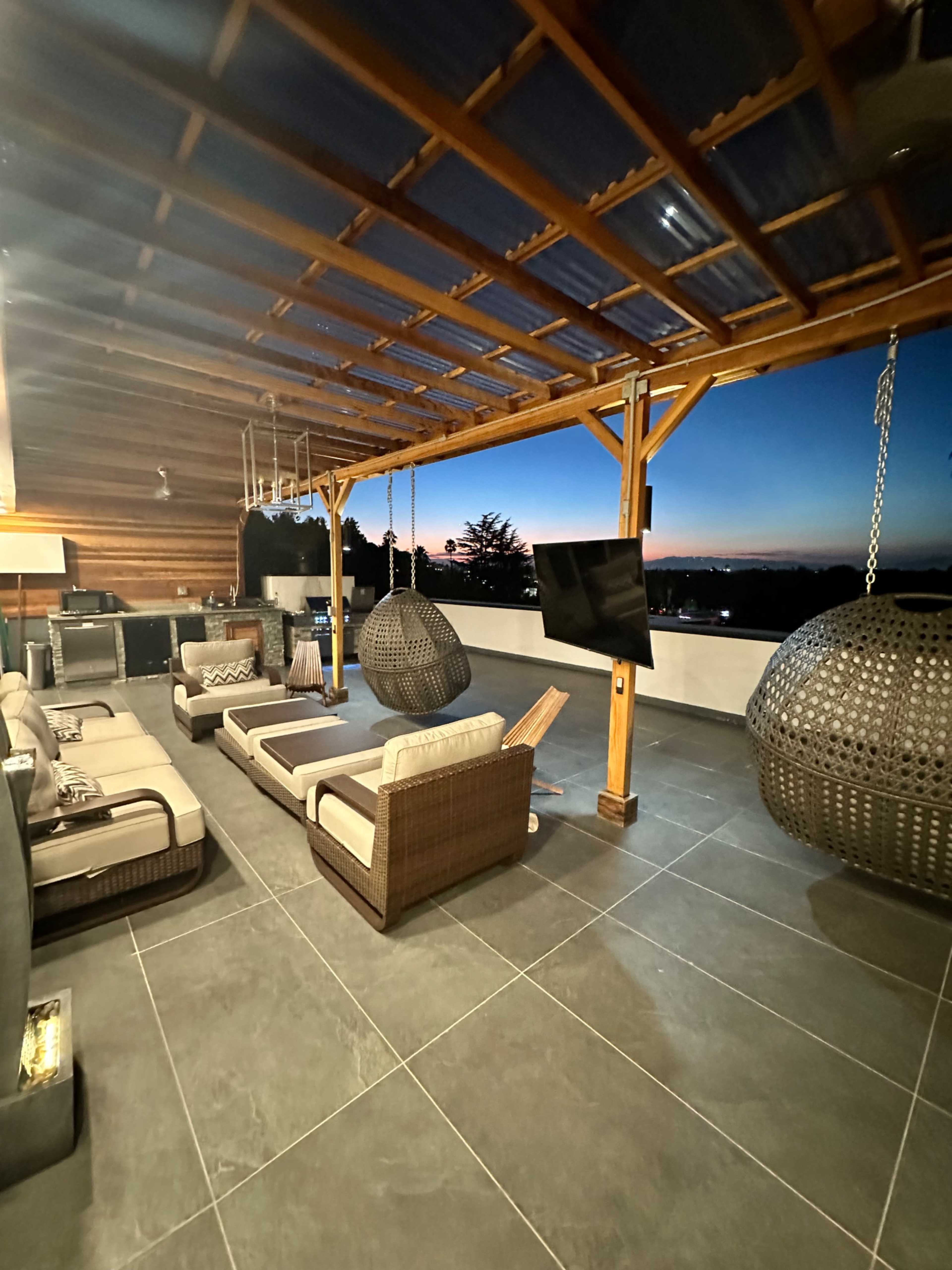 The image shows a modern outdoor lounge area with comfortable seating, hanging chairs, and a view of a twilight sky.