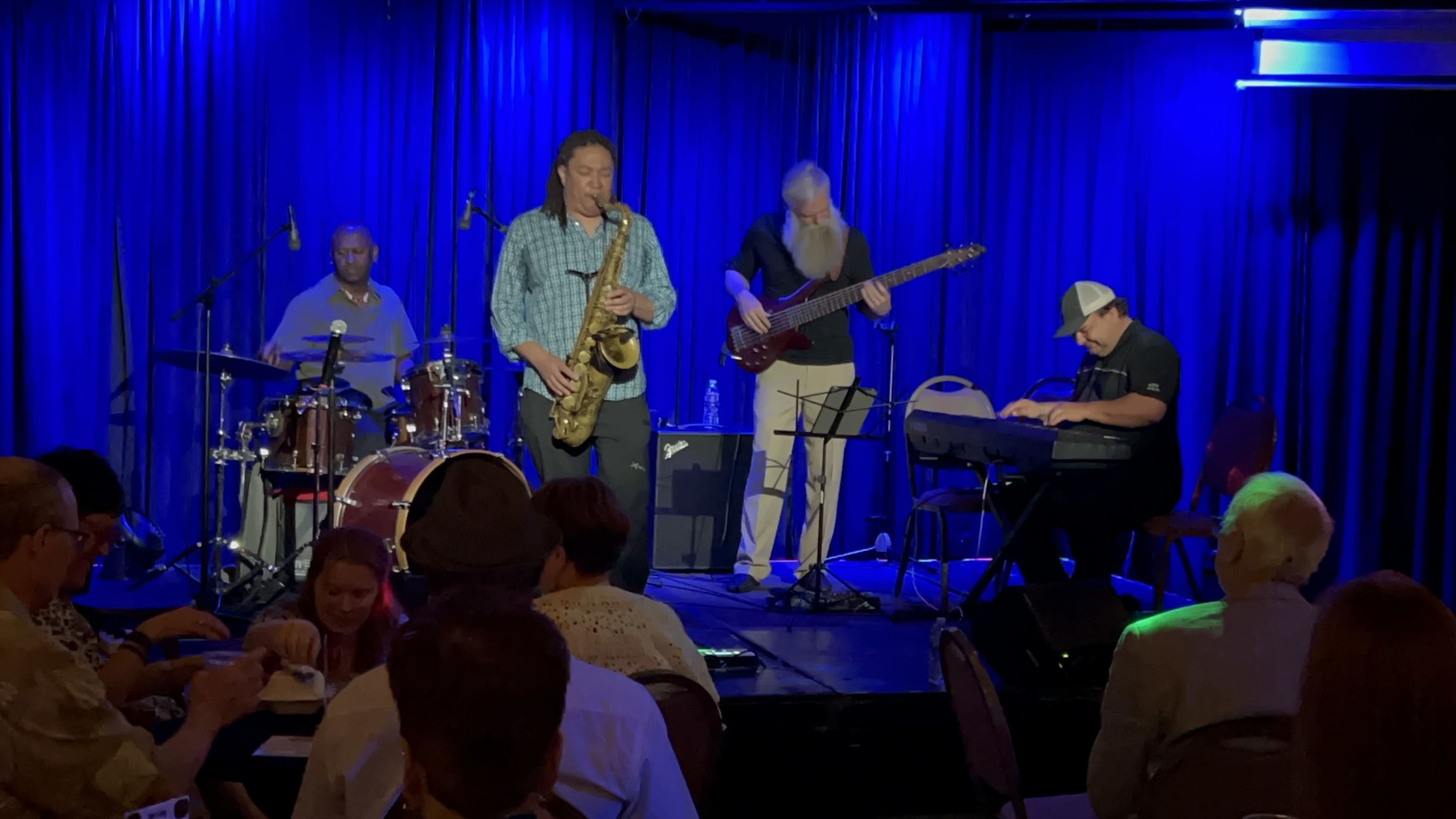 A jazz band performs on stage in front of an audience, featuring a saxophonist, drummer, bassist, and keyboardist under blue lighting.
