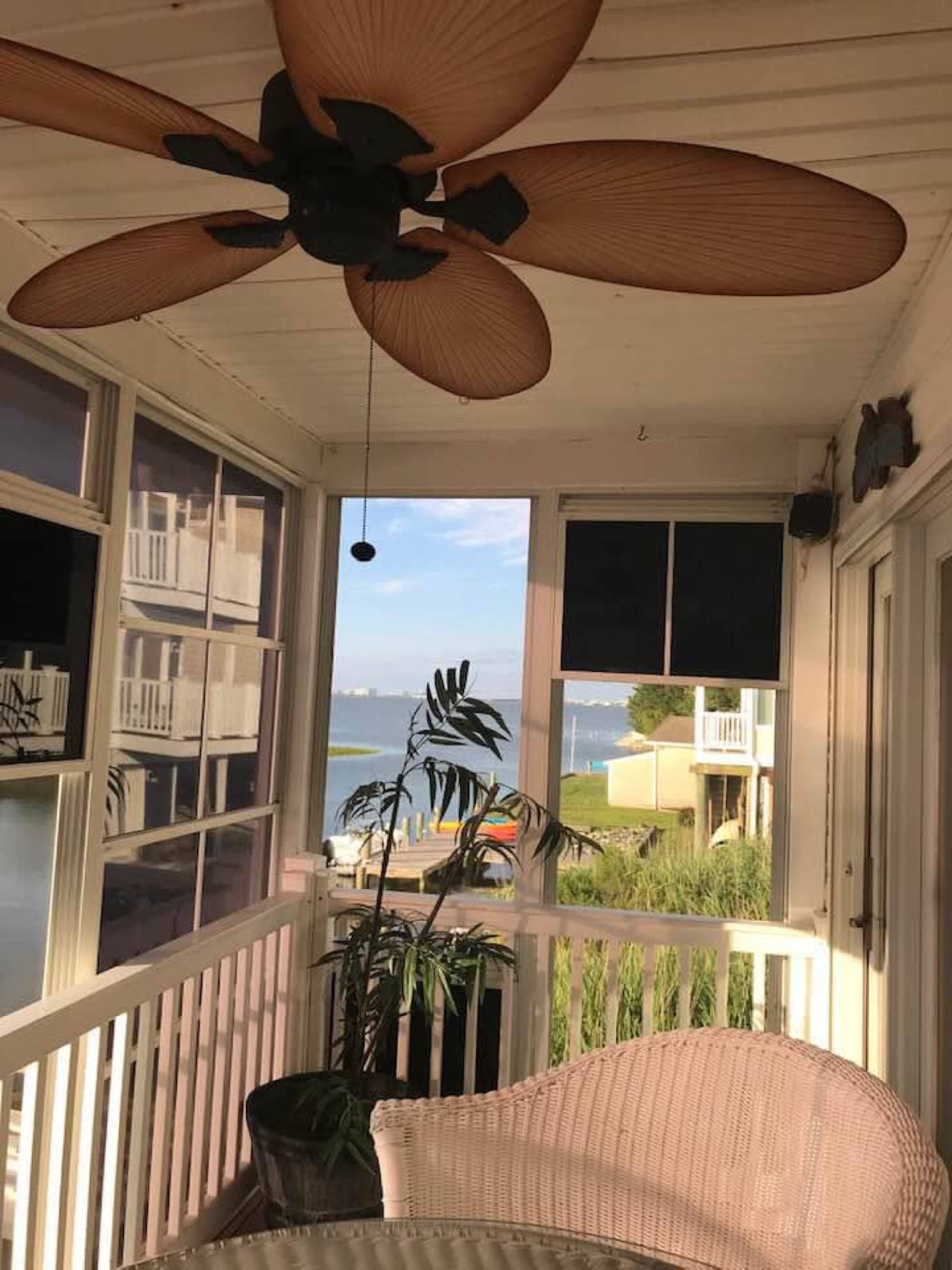 A screened porch features a ceiling fan, a potted plant, and a view of a body of water and nearby structures.