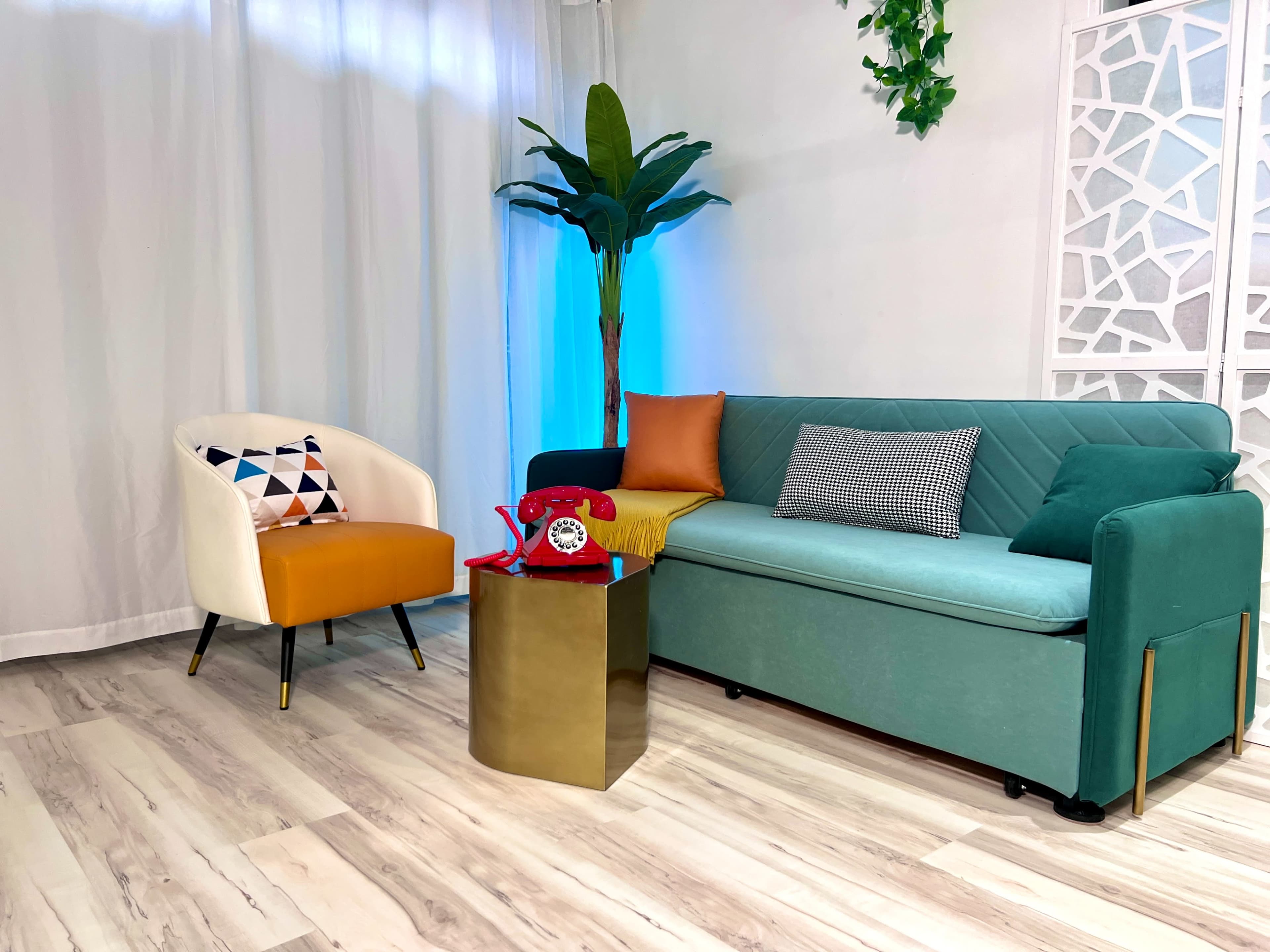 The image features a modern living space with a light-colored wooden floor, a teal sofa, an orange and white accent chair, and a gold side table, complemented by artificial plants and soft lighting.