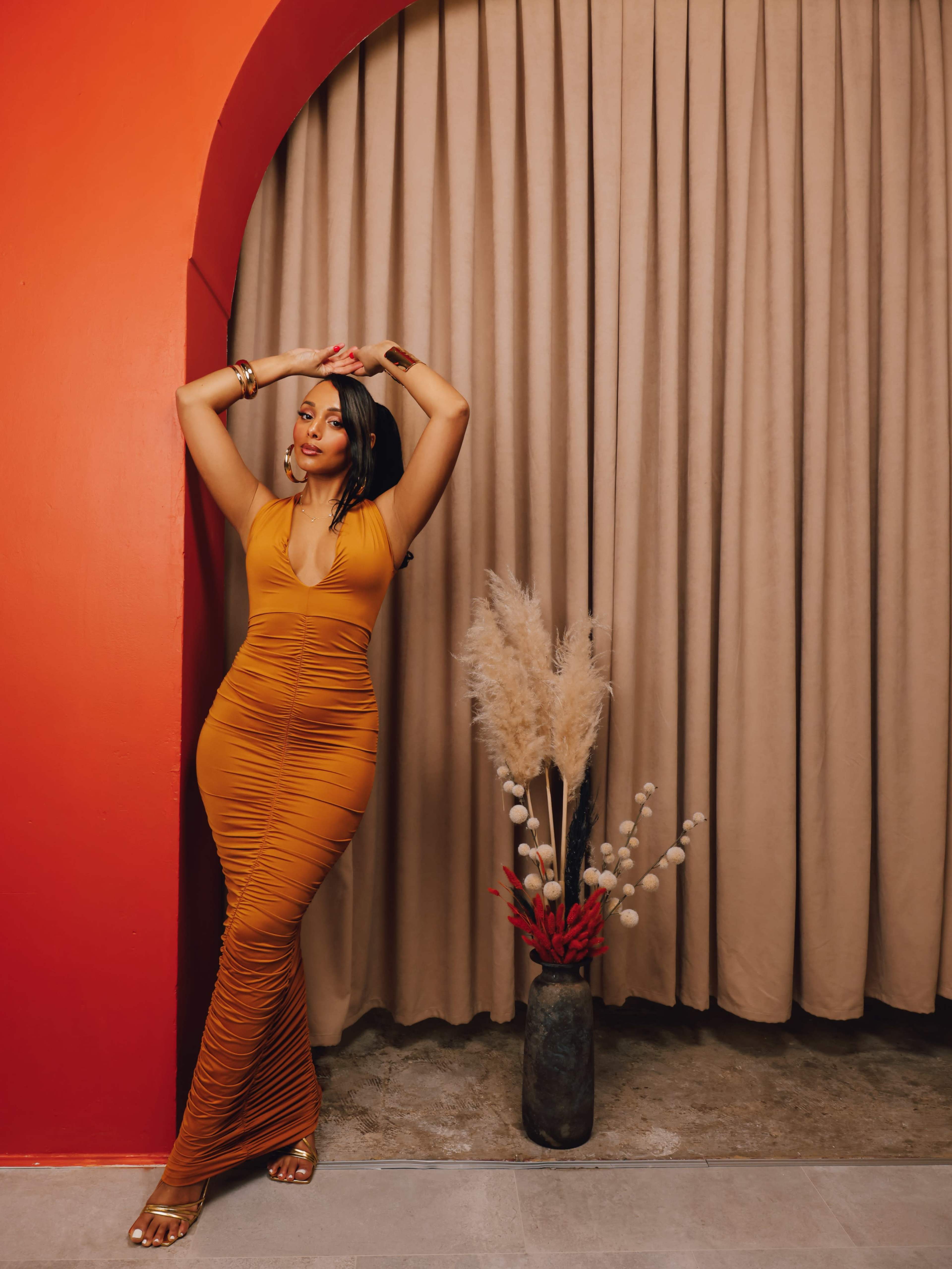 A woman wearing a form-fitting, orange dress poses beside a neutral-colored curtain and a decorative vase with dried flowers on the floor.