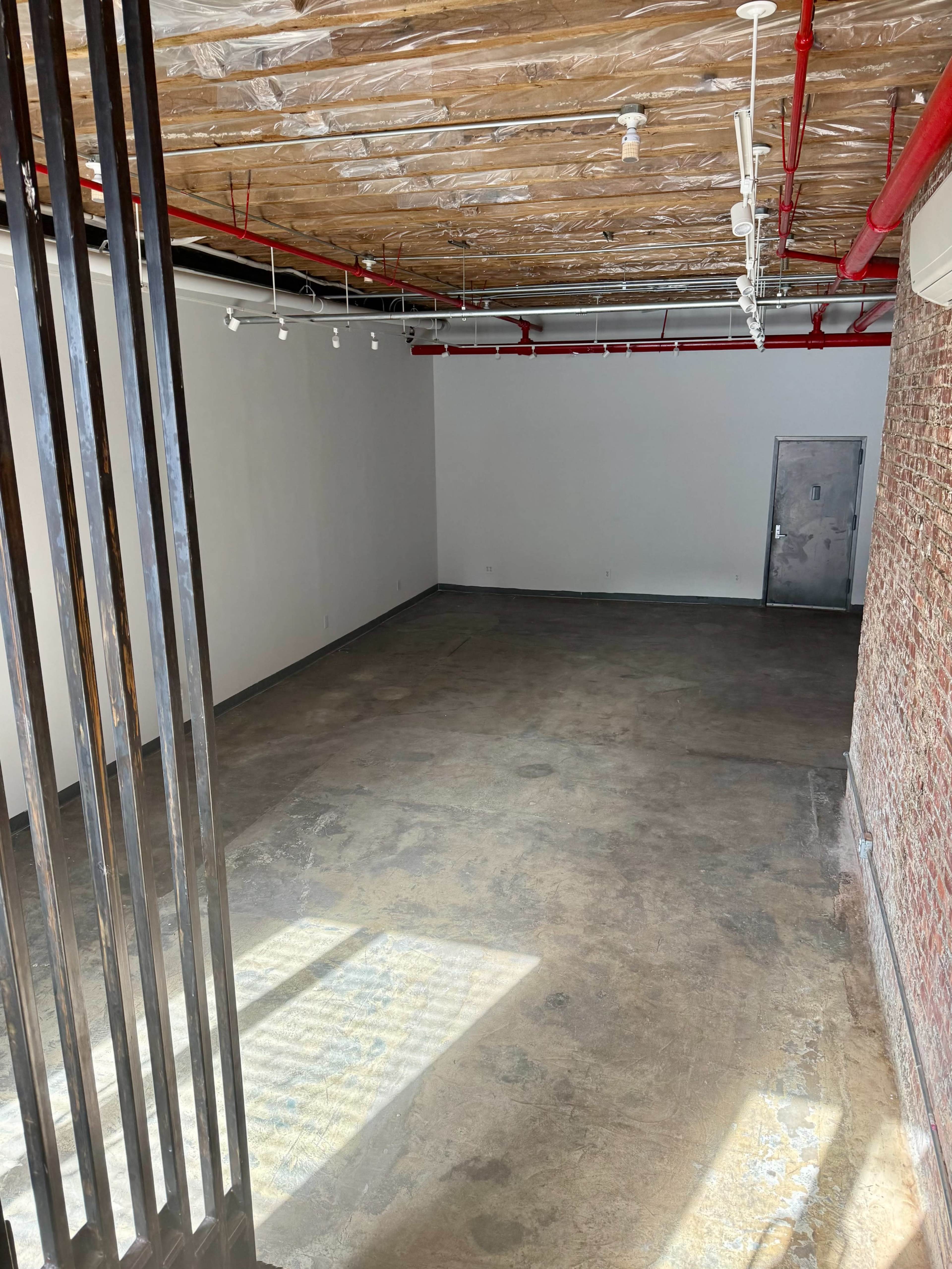 The image shows a spacious, empty interior room with exposed brick walls, a concrete floor, and a ceiling featuring visible pipes and lights.