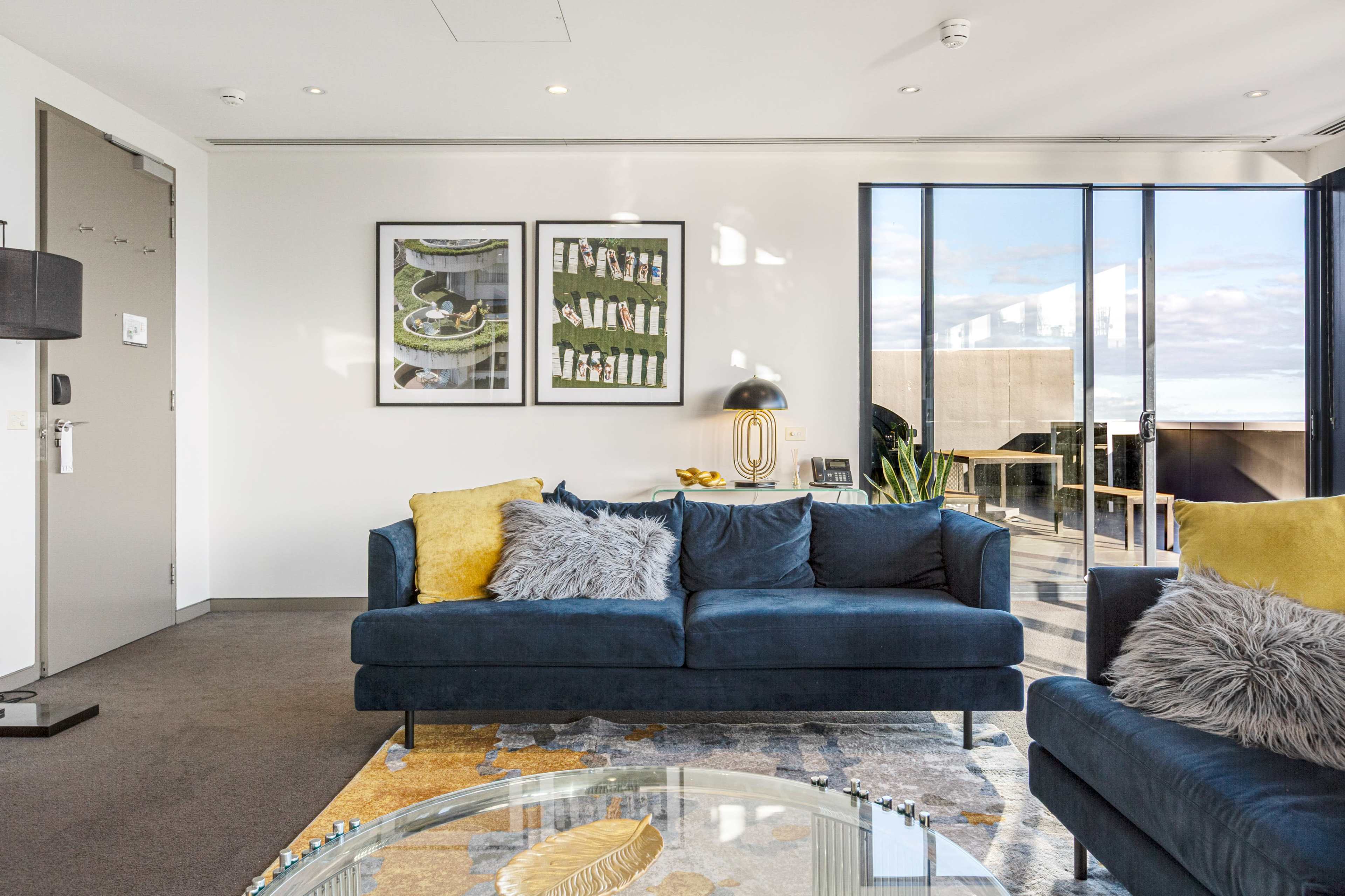 The image shows a modern living room featuring two blue sofas with decorative cushions, a glass coffee table, and large windows allowing natural light to enter the space.