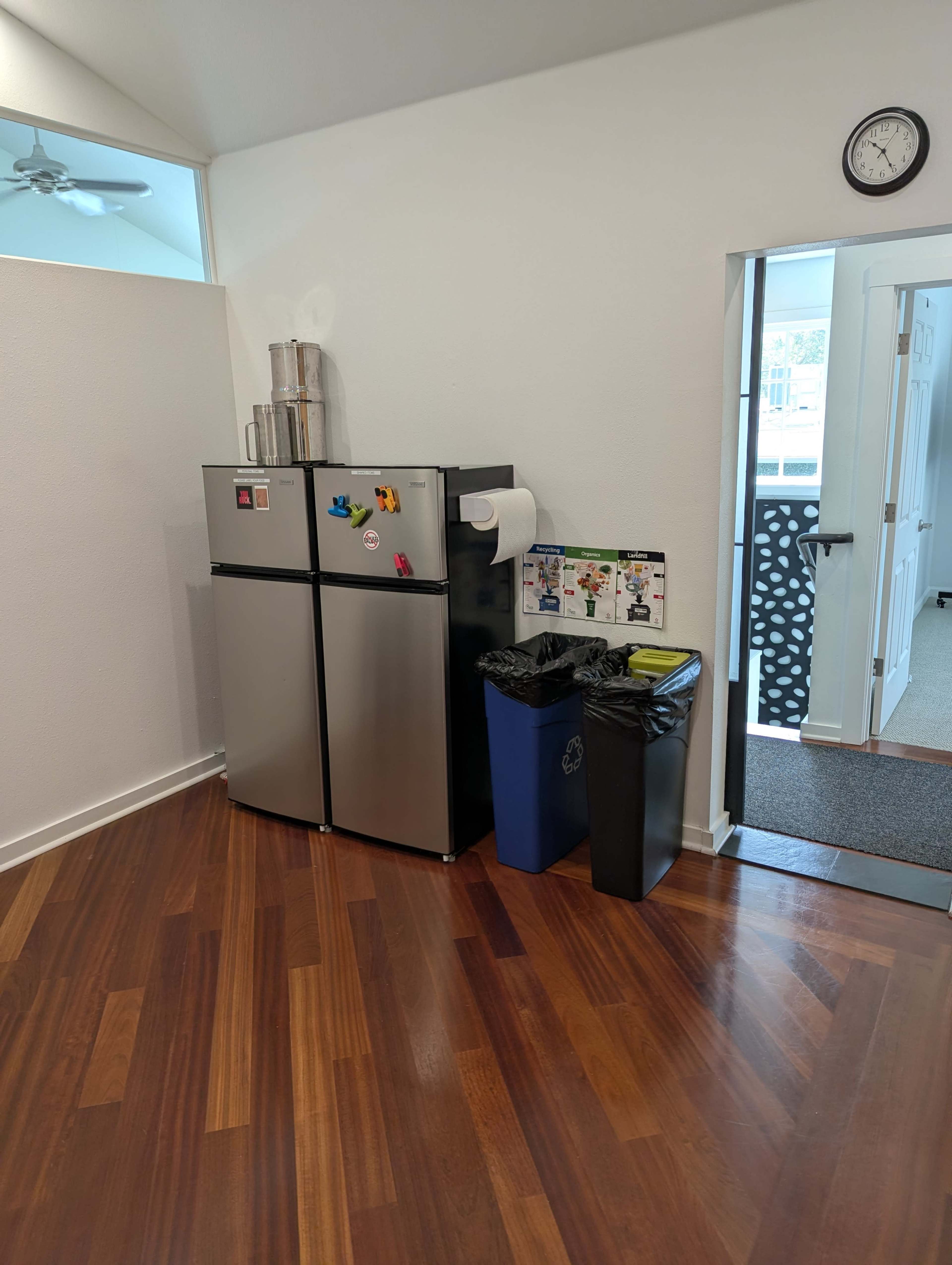 The image shows a kitchen area featuring a silver refrigerator, a trash can, a recycling bin, and a wall clock, with wooden flooring and a doorway in the background.