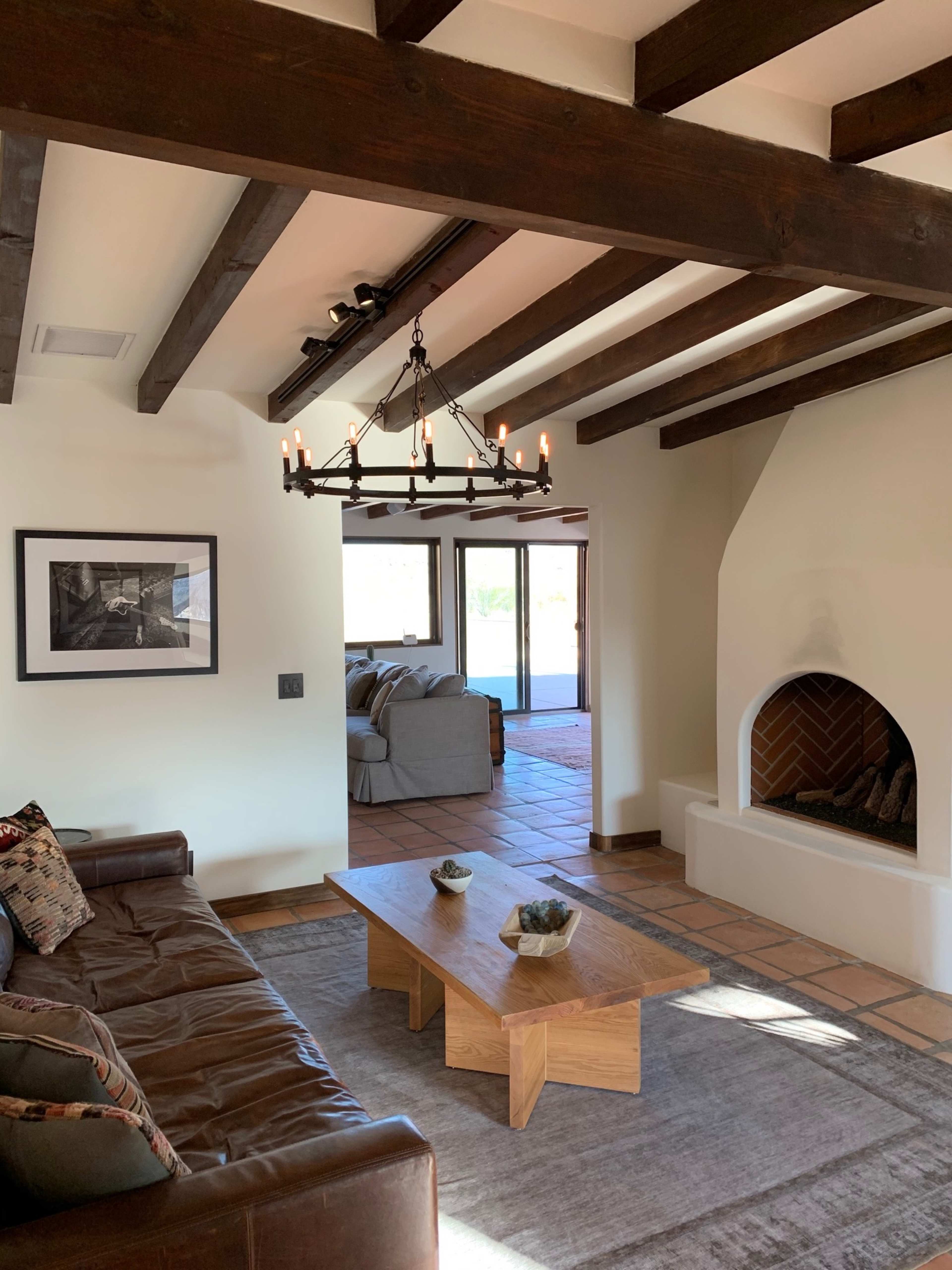 The image shows a living room with a leather couch, wooden beams on the ceiling, a chandelier, and a fireplace, alongside a coffee table and a view of outdoor space through large windows.