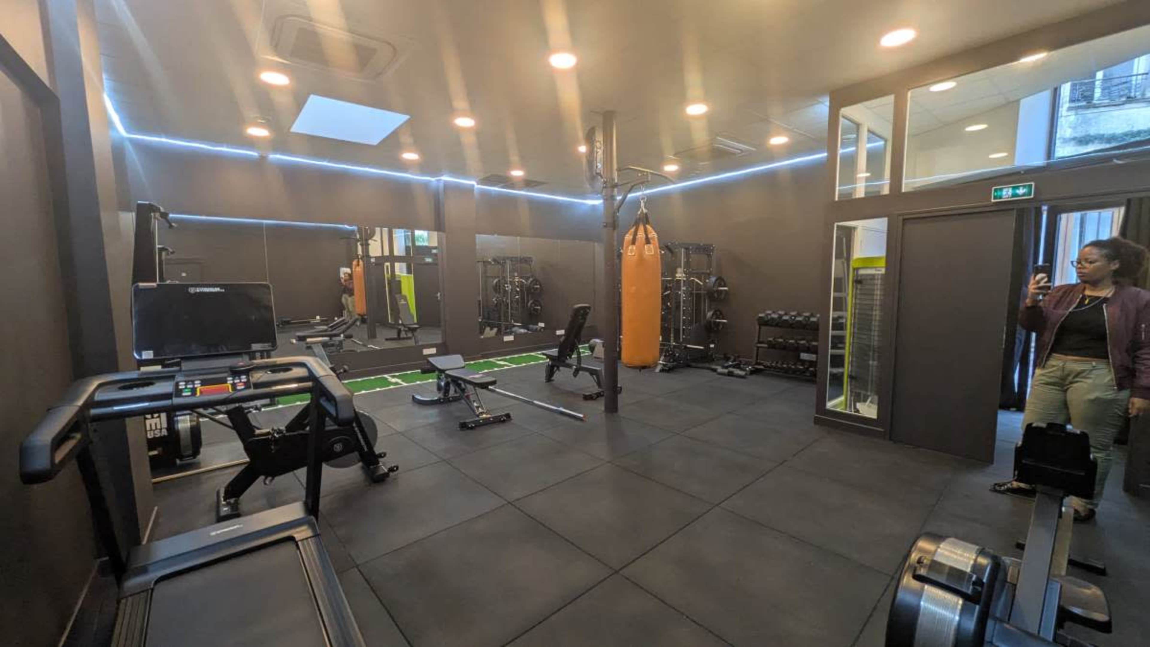 The image shows a modern gym interior equipped with various exercise machines, free weights, and a punching bag, with a person standing near the entrance.