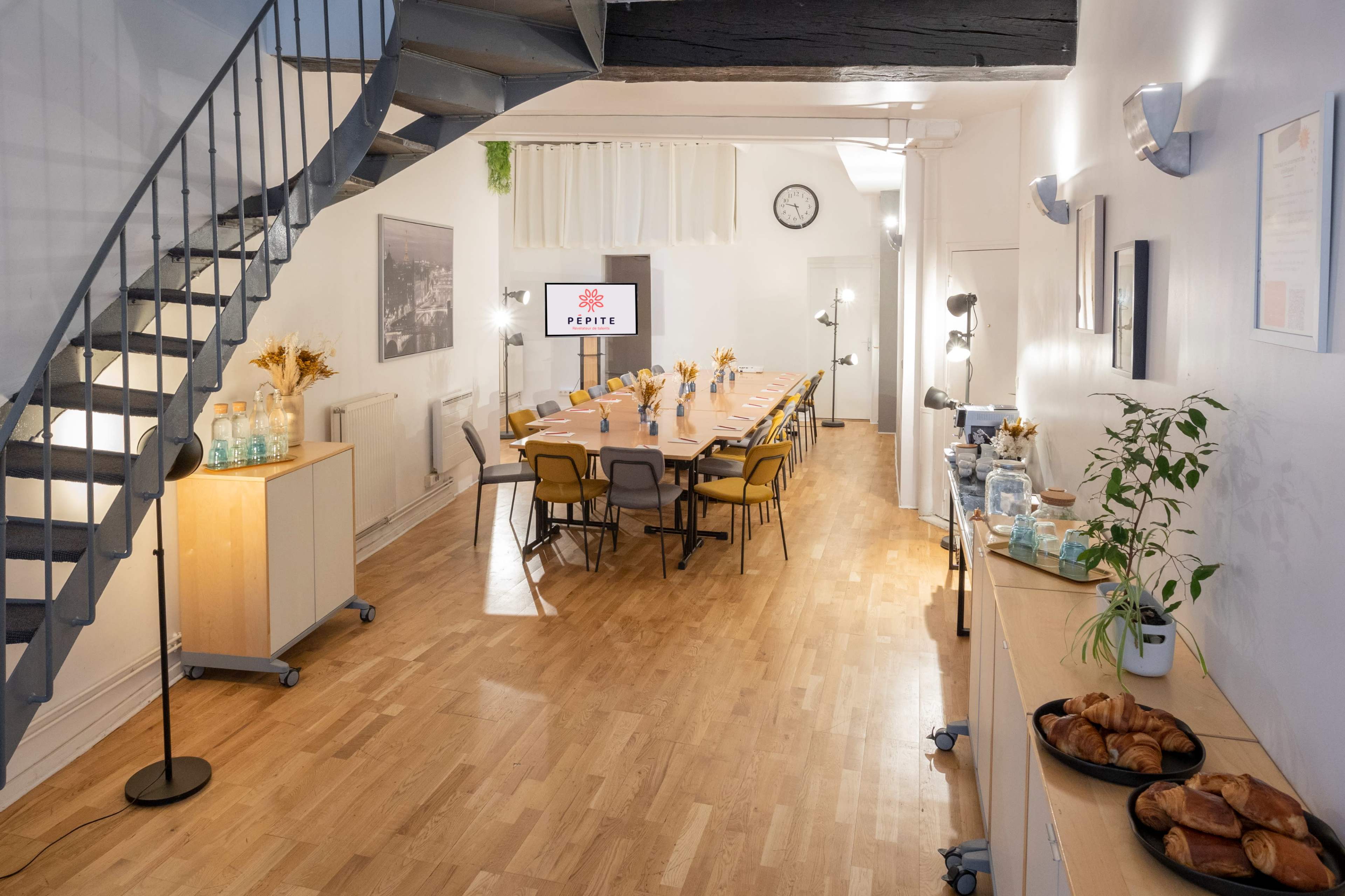 A well-lit, modern meeting room with a long table set for an event, featuring pastries and beverages along the walls, and a staircase in the background.