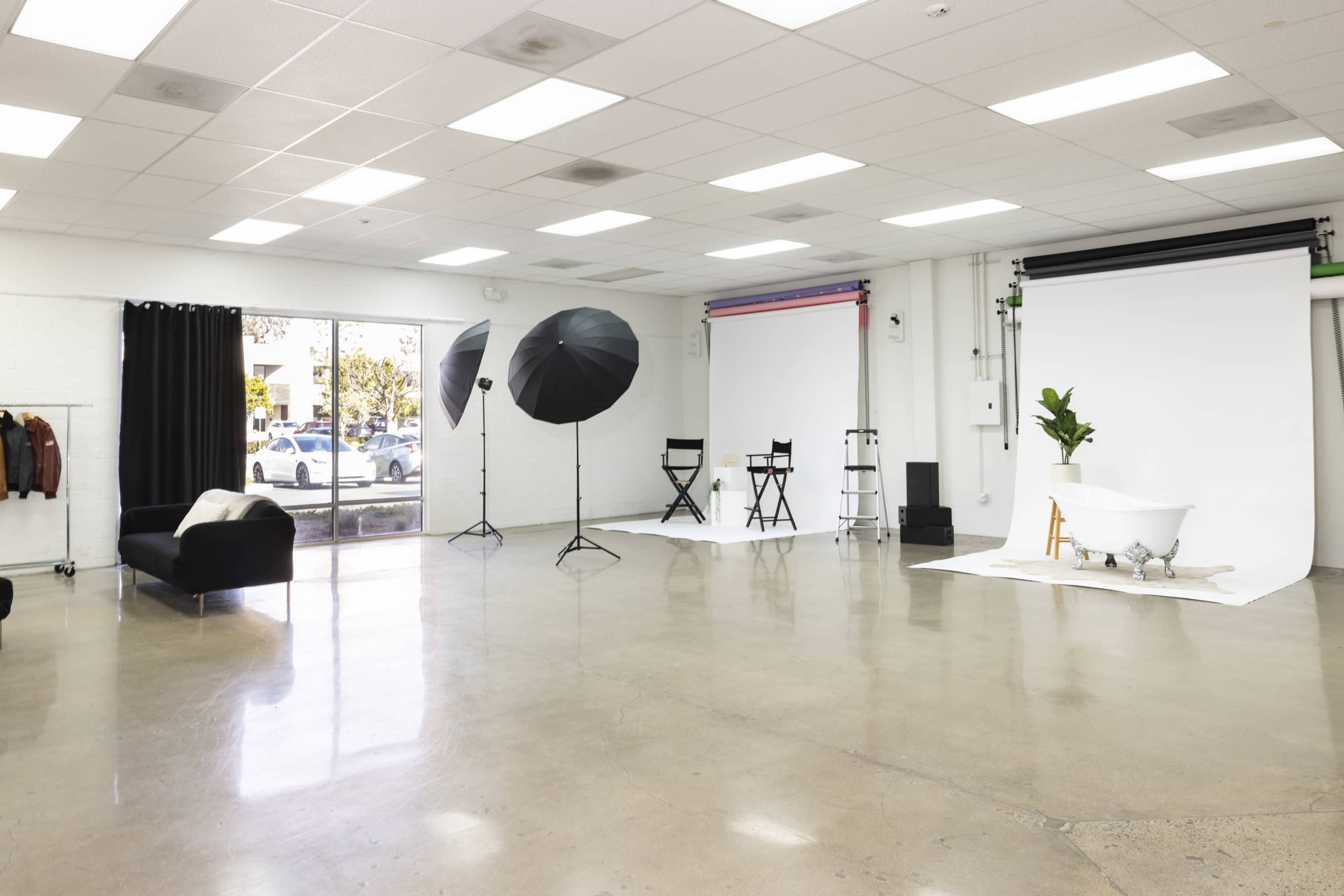 A spacious photography studio featuring a black couch, two director's chairs, portable light umbrellas, a vintage bathtub, and a large backdrop setup.