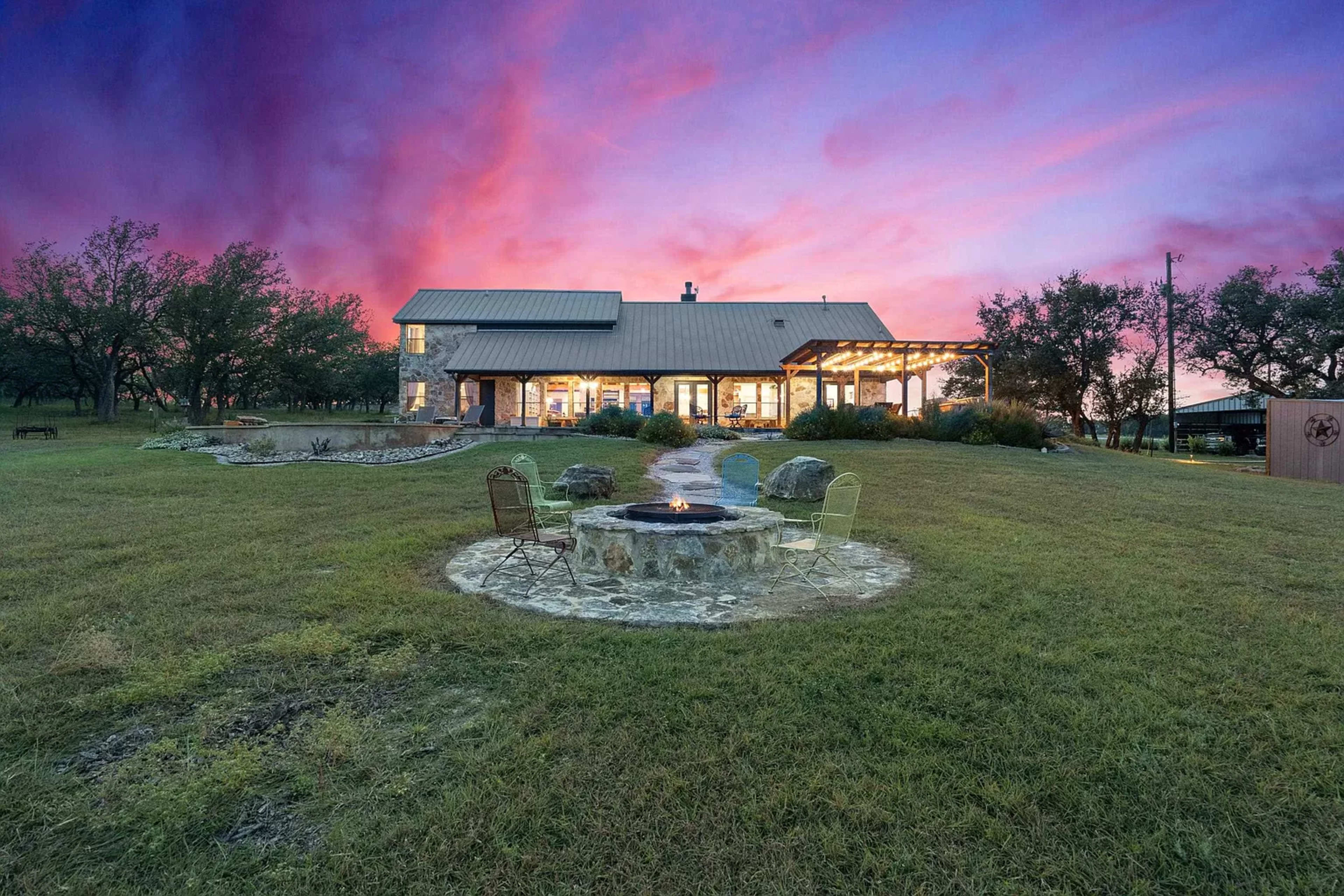 A large house with a metal roof, surrounded by trees, features a stone fire pit and chairs in the foreground under a colorful sunset sky.