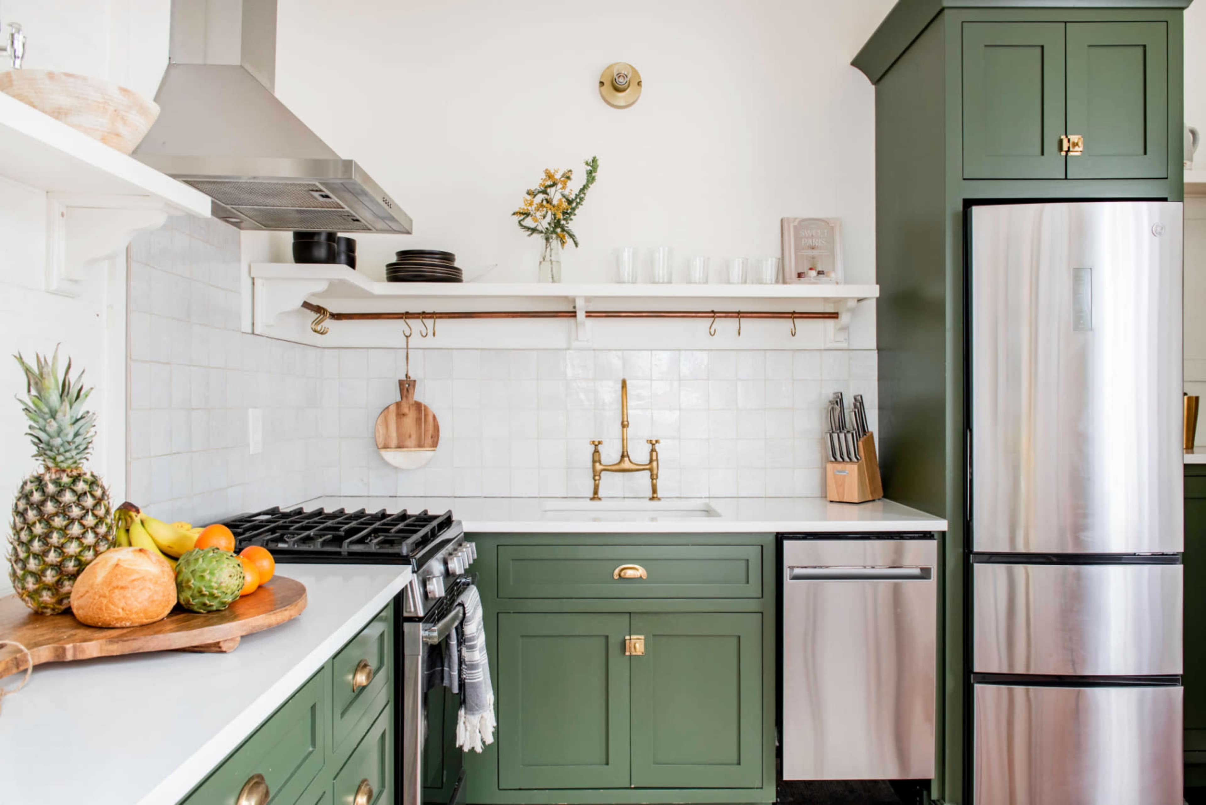 The image depicts a modern kitchen featuring green cabinetry, a stainless steel refrigerator, a gas stove, and a countertop adorned with various fruits.