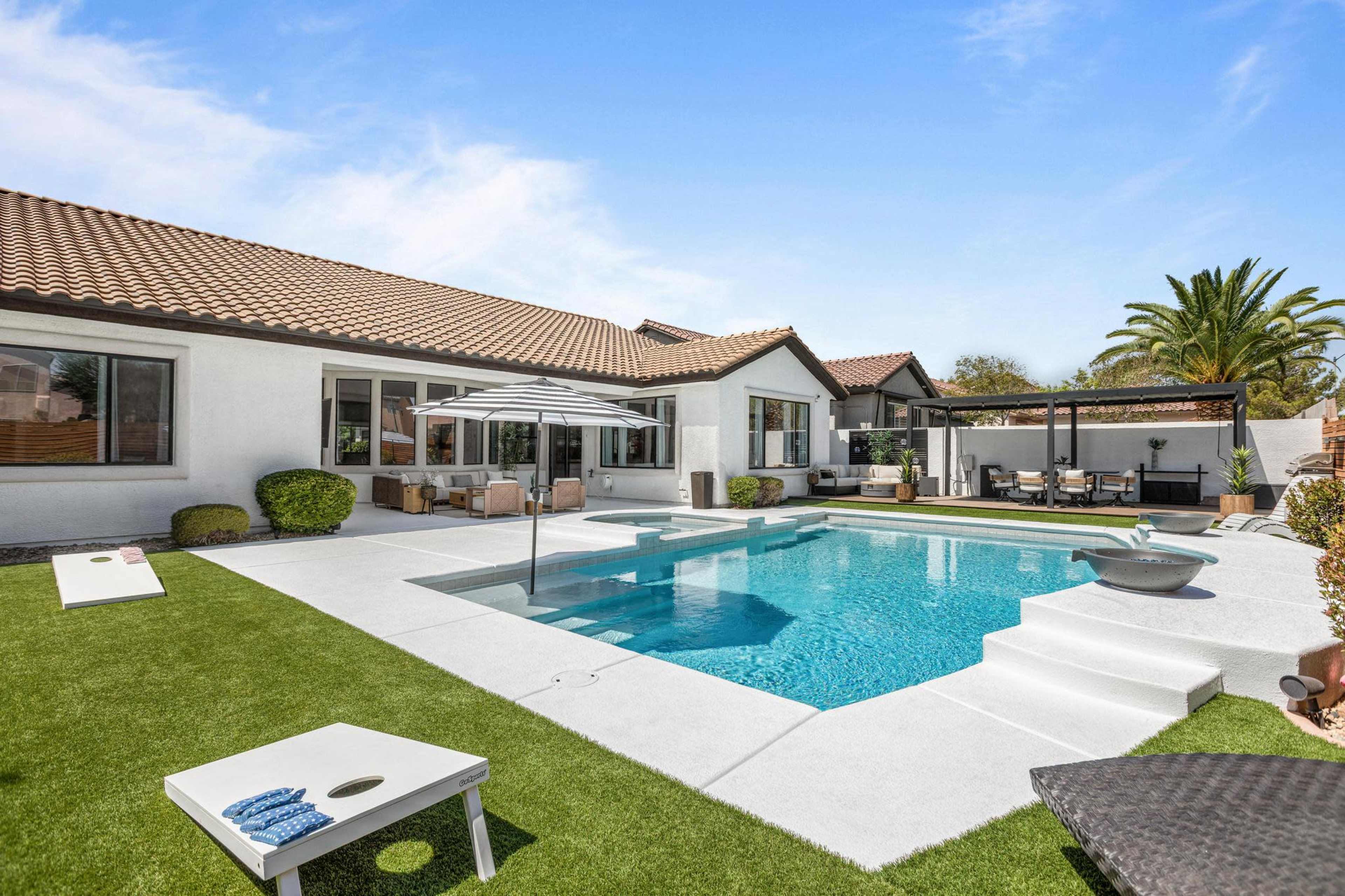 The image shows a modern backyard featuring a swimming pool, lounge chairs, a shaded seating area, and a neatly landscaped lawn.
