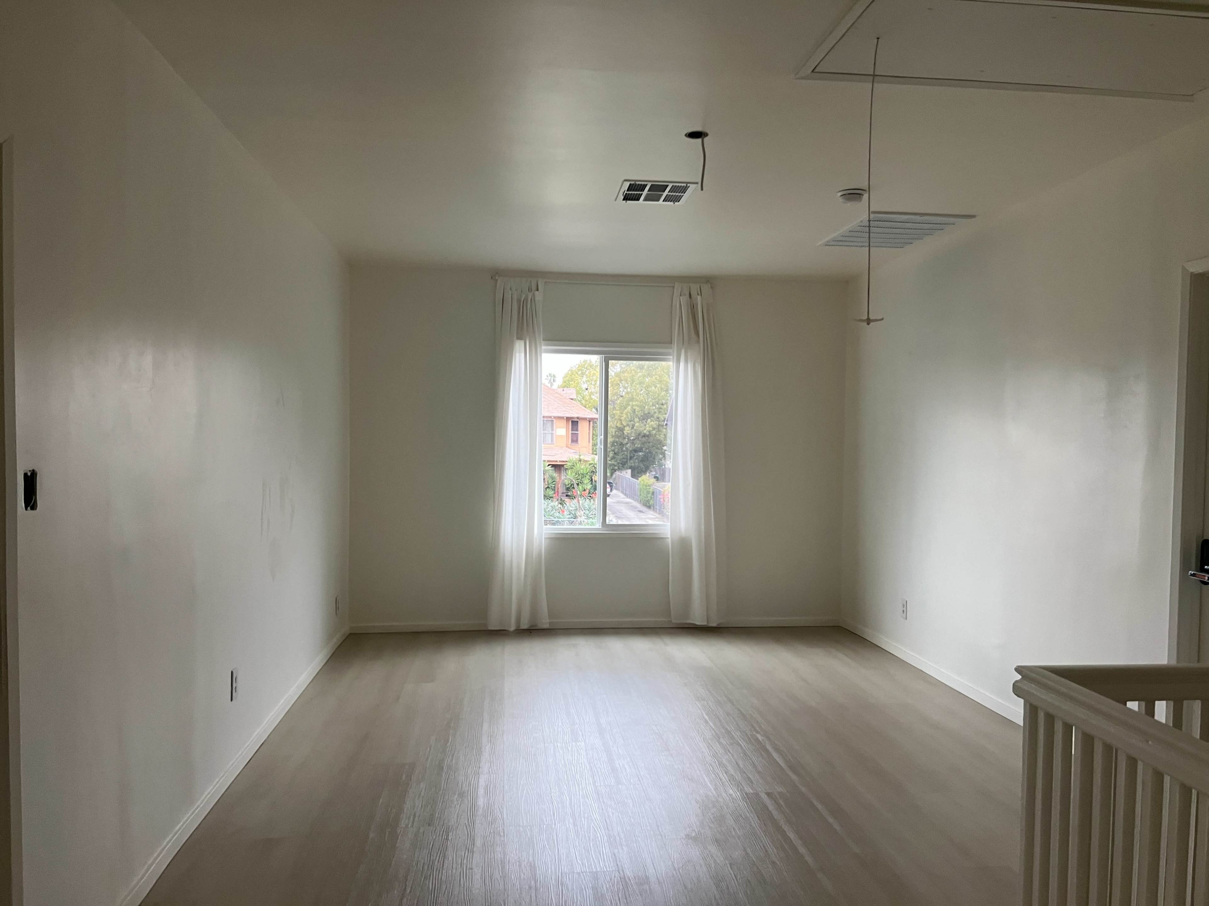 The image shows a minimally furnished room with white walls, light wooden flooring, and a window covered by sheer curtains.
