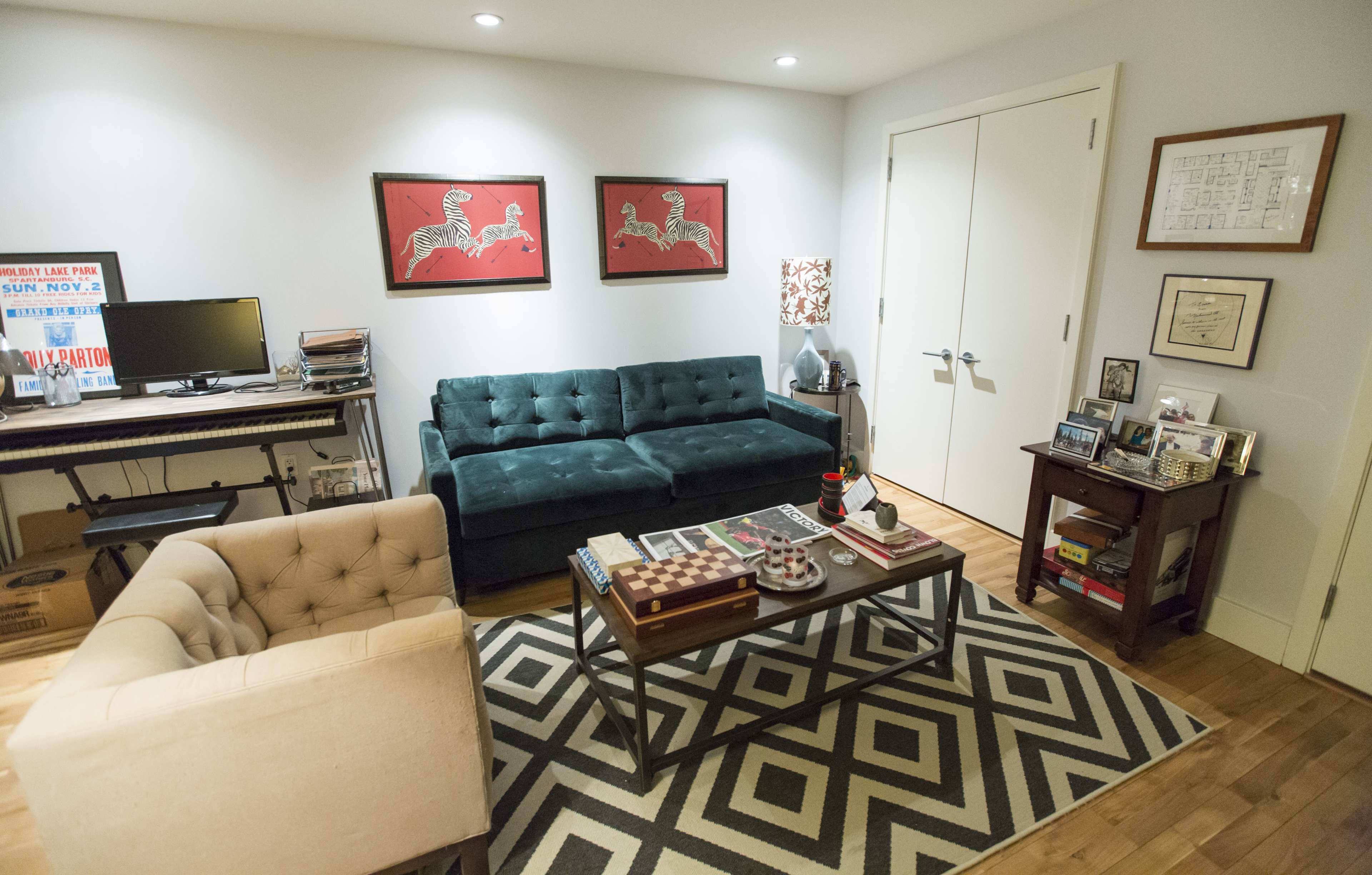 A small living room with a green tufted couch, a beige armchair, a coffee table with books and a decorative item, and two red artwork pieces on the wall.