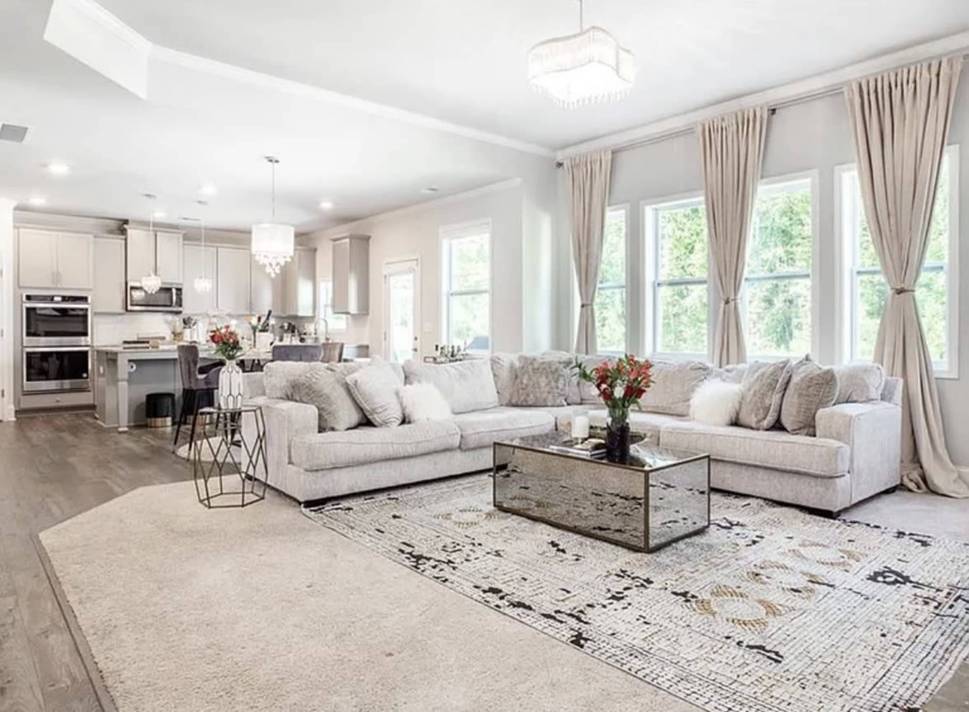 The image depicts a modern living room featuring a large, light-colored sectional sofa, a glass coffee table, and a patterned area rug, with large windows allowing natural light to fill the space.