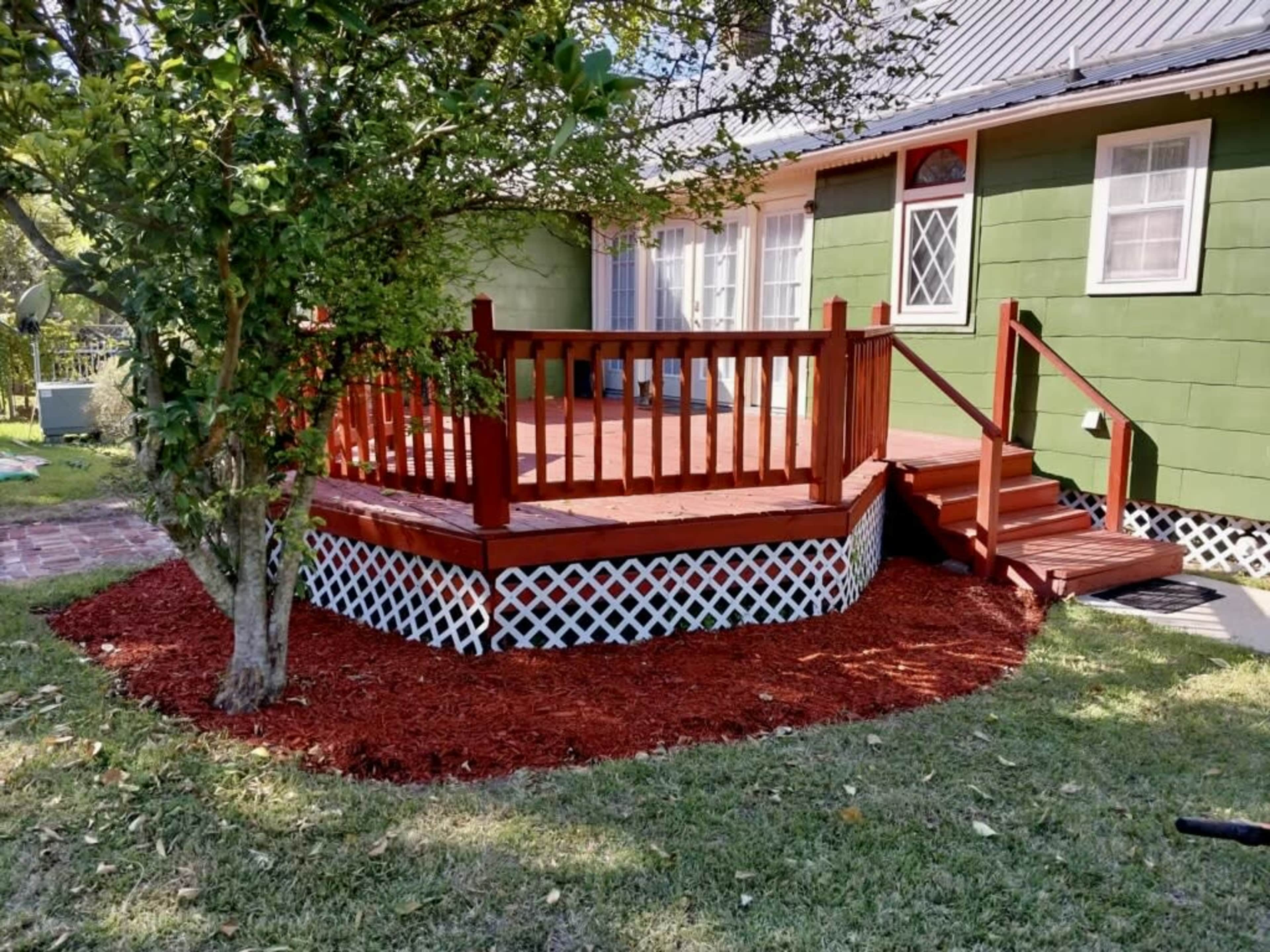 The image shows a wooden deck with a red railing and staircase, surrounded by freshly laid red mulch, next to a green house.