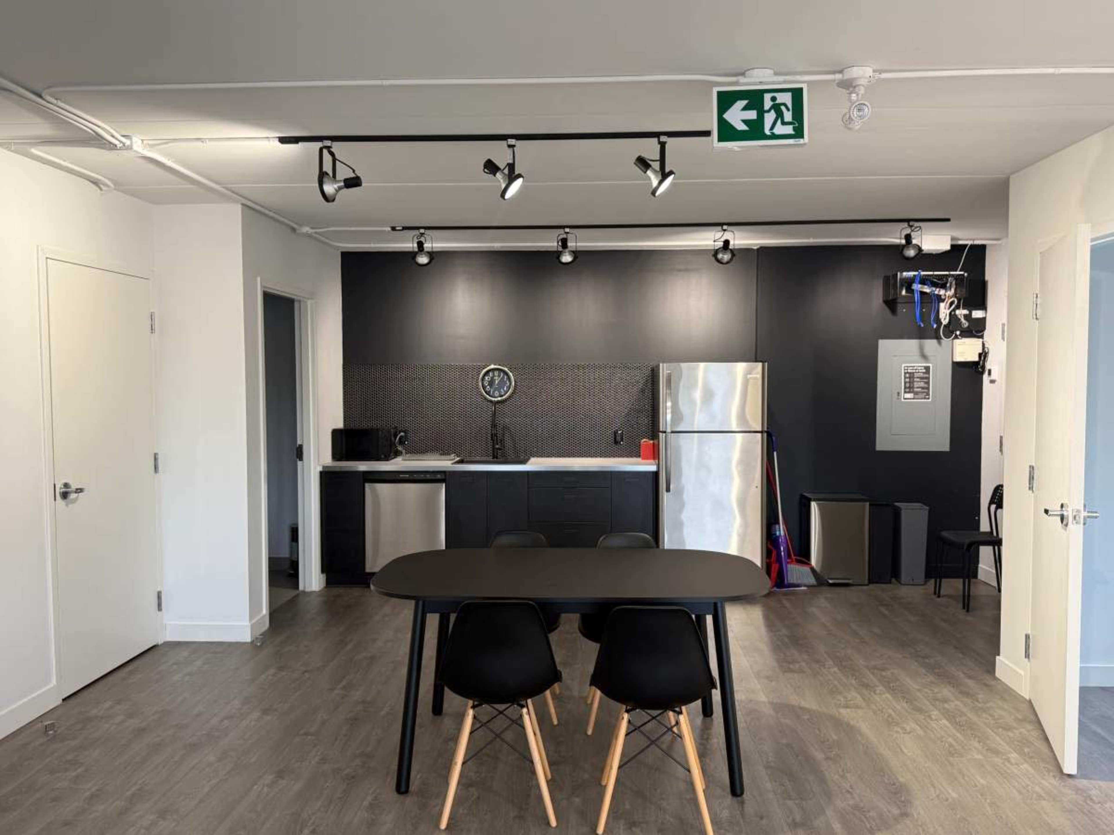 The image shows a modern, open-concept kitchen and dining area featuring a black wall with a clock, stainless steel appliances, and a simple dining table with black chairs.