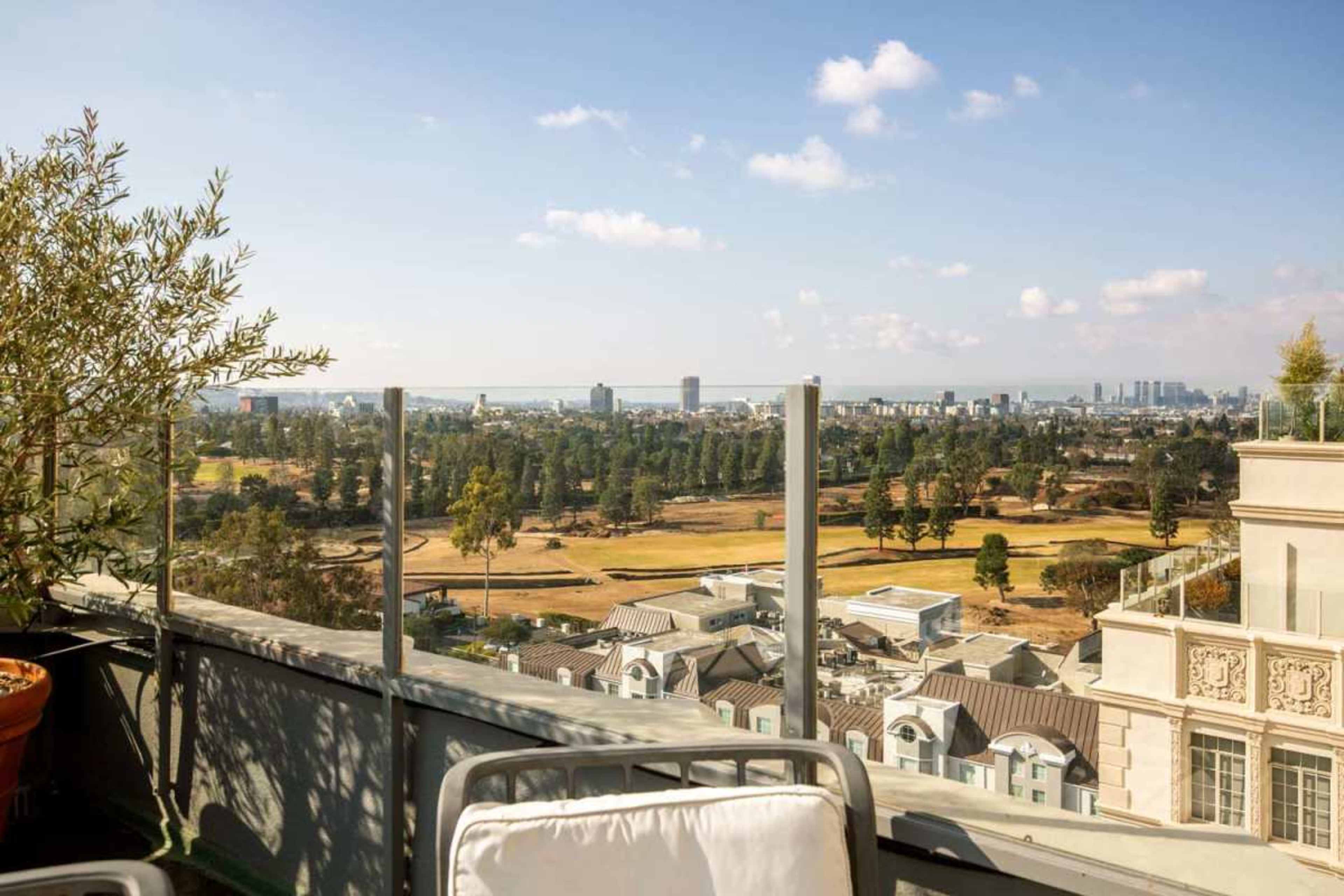 The image shows a rooftop view overlooking a cityscape with a golf course and distant skyscrapers under a clear blue sky.