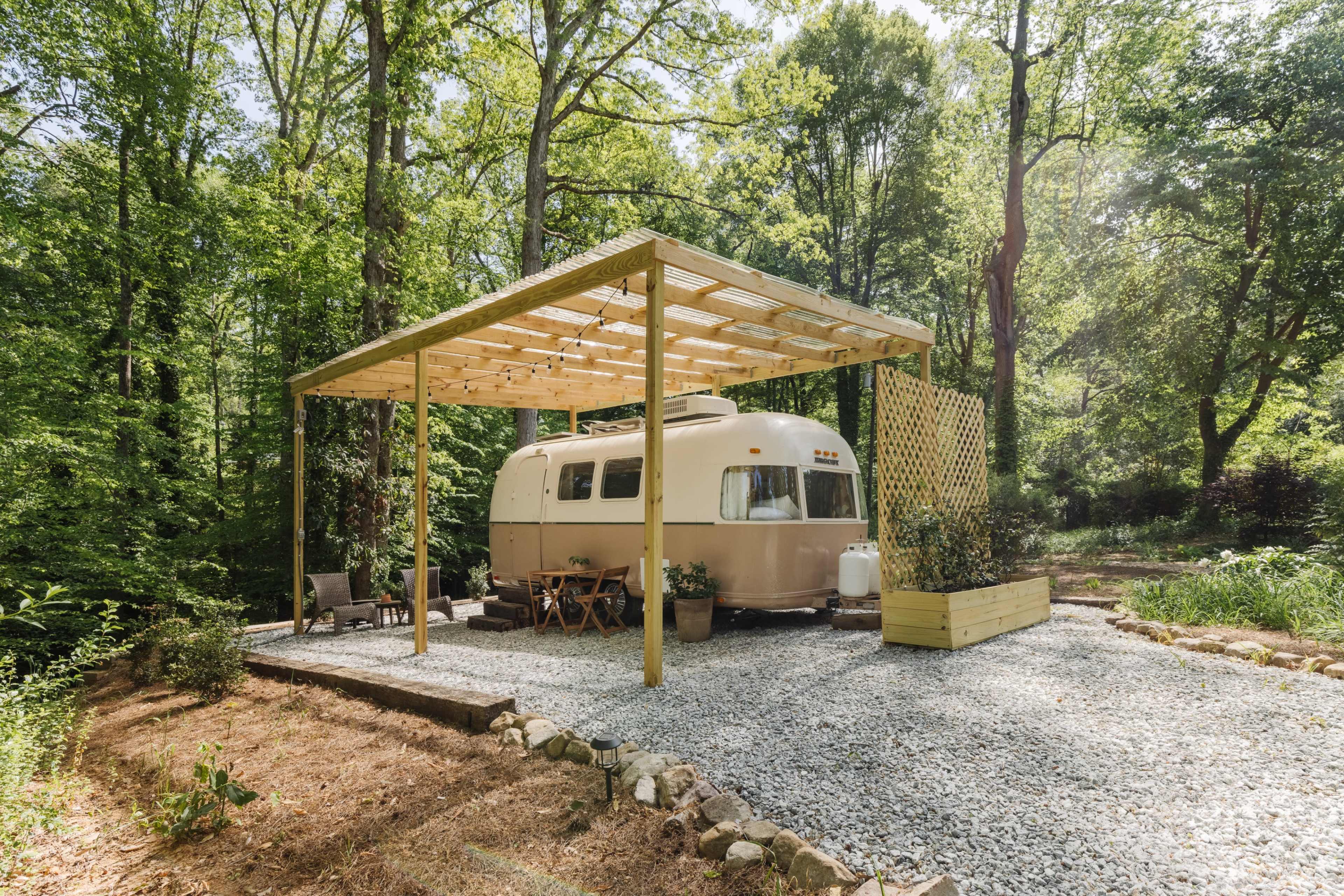 A vintage Airstream trailer is parked underneath a wooden pergola in a wooded clearing, surrounded by gravel and seating areas.