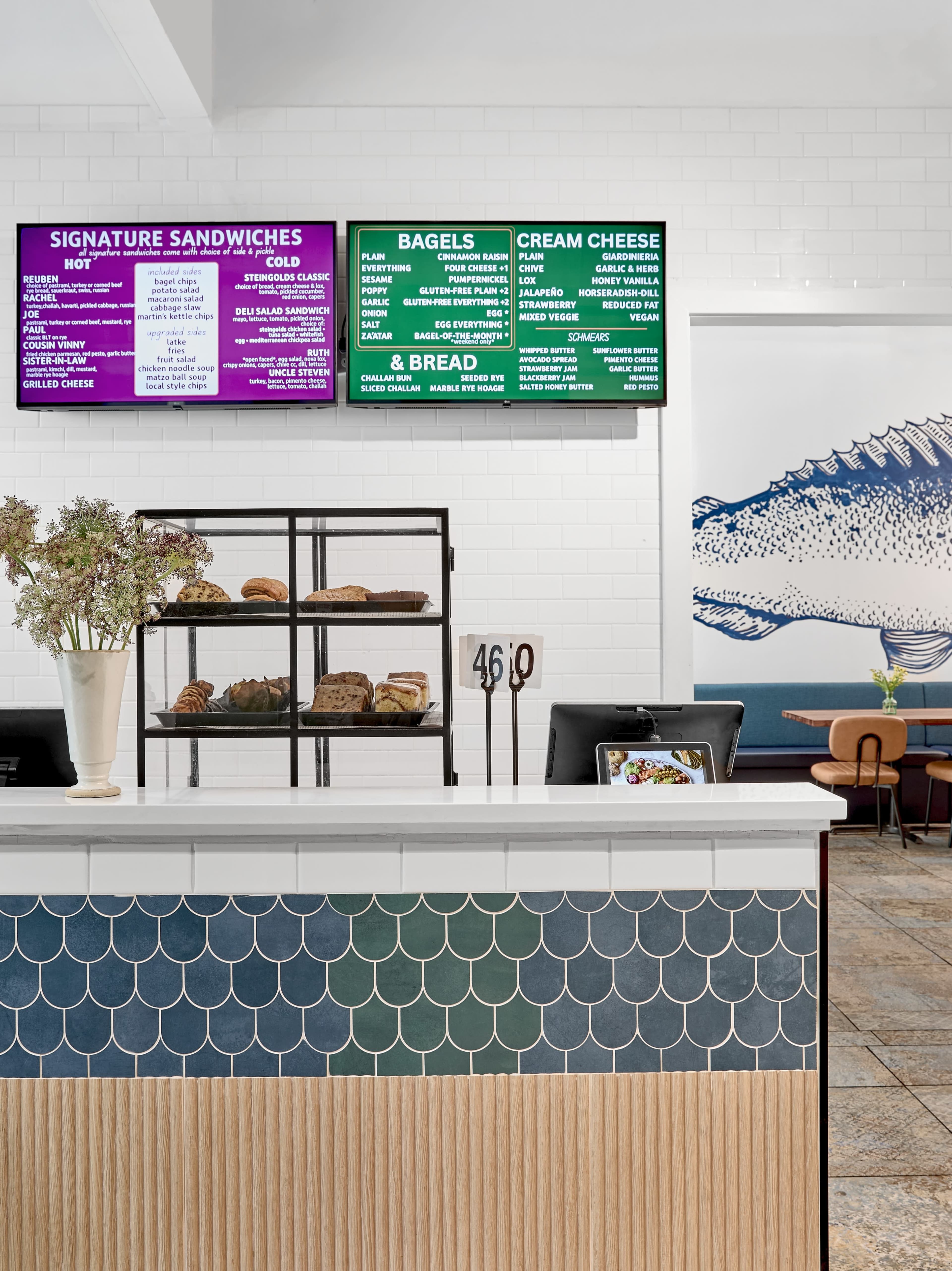 A bright café interior features a display case with baked goods, a menu board listing various food items, and a decorative wall art of a fish.