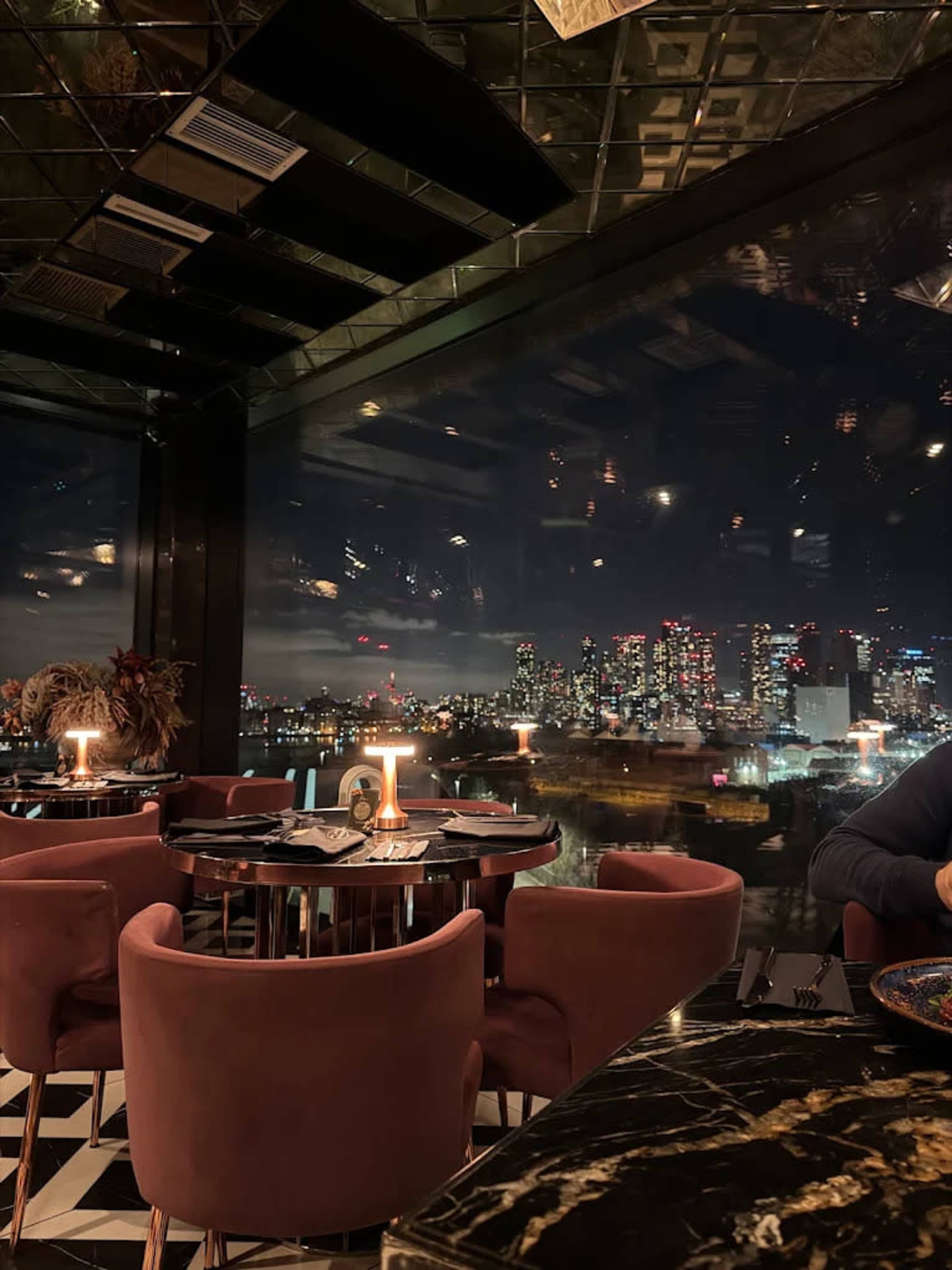 The image shows an upscale restaurant with elegant seating, dark walls, and large windows revealing a city skyline at night.