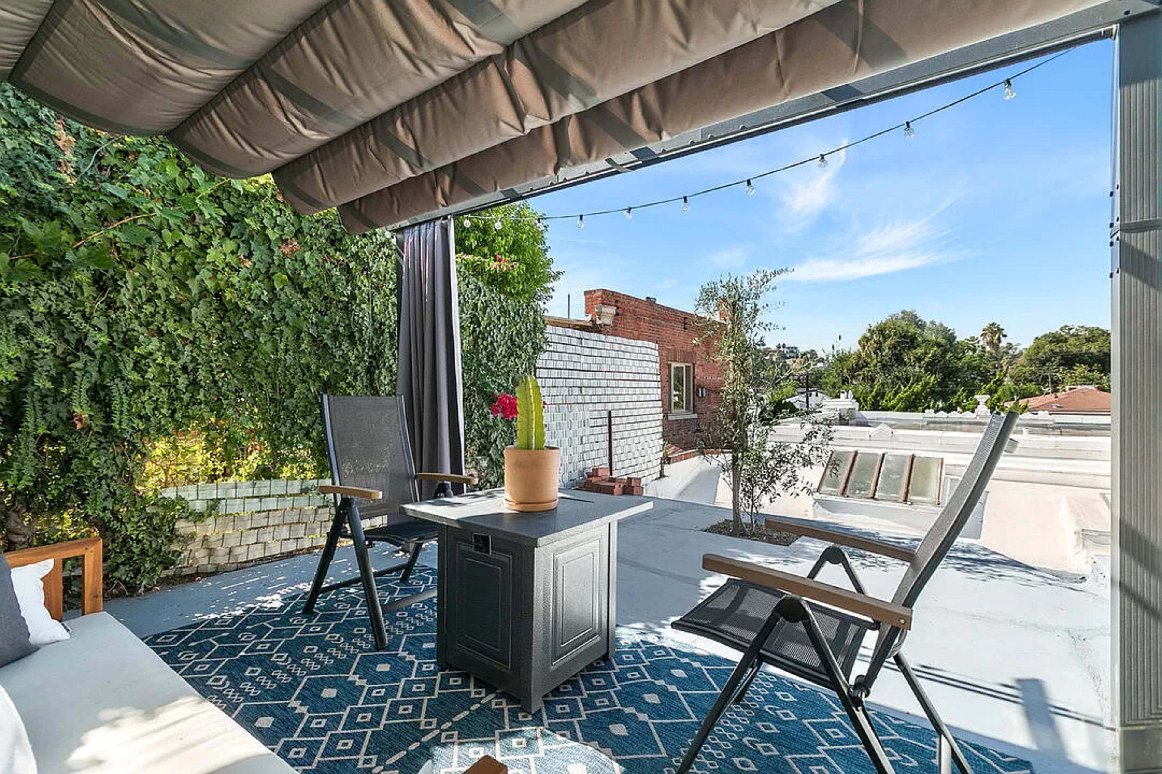 A rooftop patio features a small table with a plant, two chairs, and a seating area, surrounded by greenery and a clear blue sky.