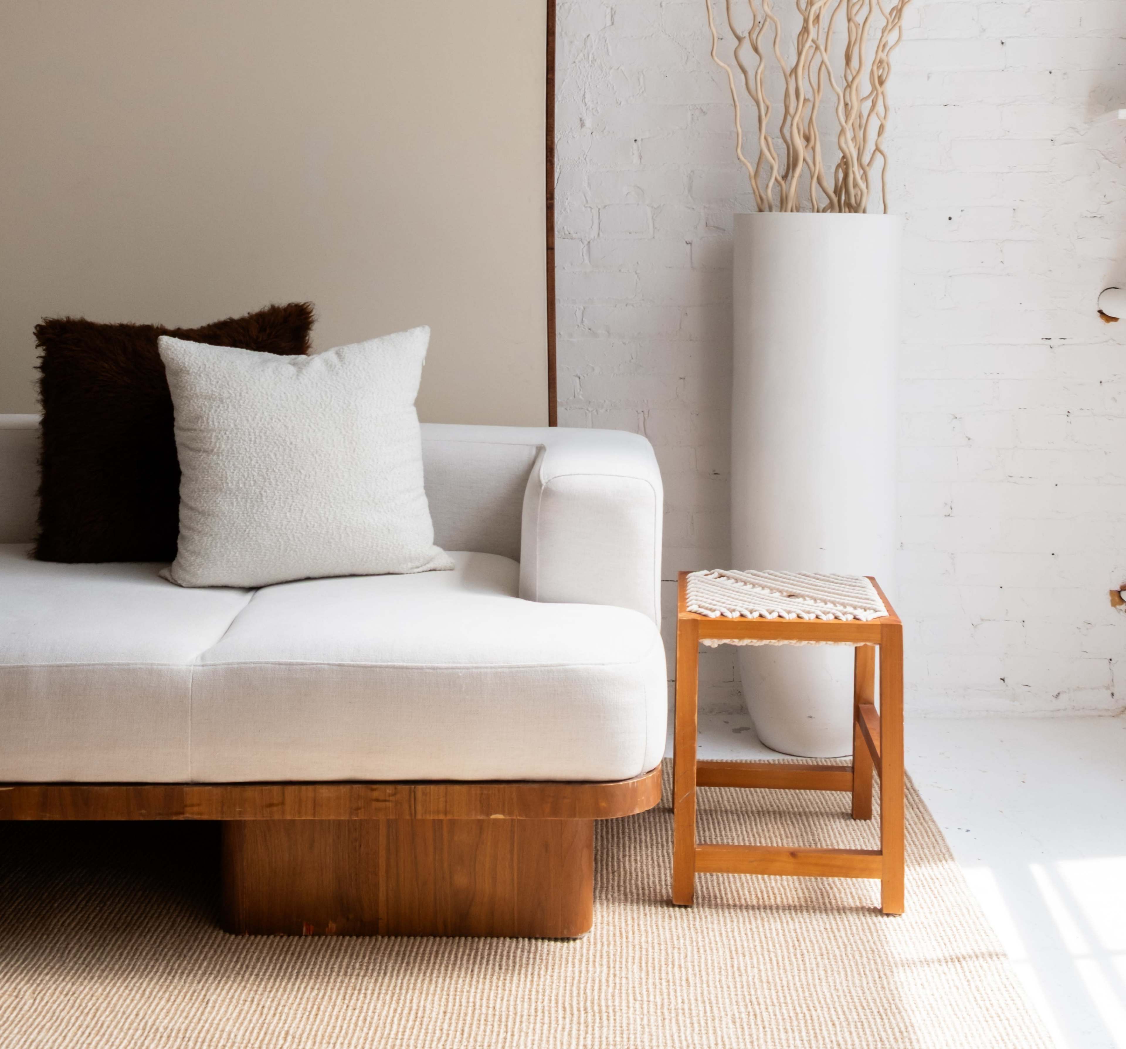 The image shows a light-colored sofa with a decorative pillow next to a wooden side table and a tall white vase with dried branches, all positioned in a bright, minimalist interior.