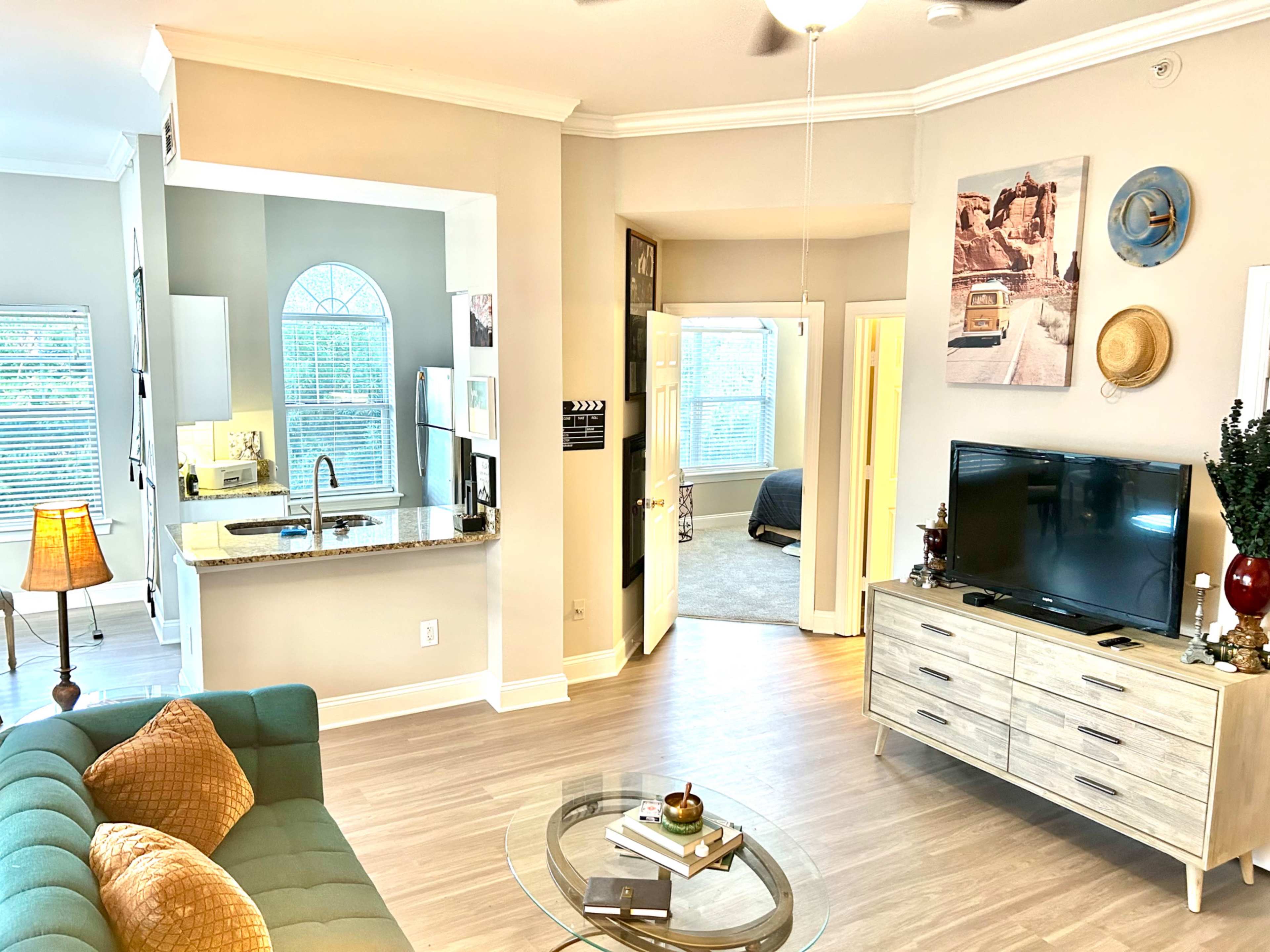 The image shows a cozy living space featuring a green sofa, a round glass coffee table, and a television, with an open view into a kitchen and a doorway leading to a bedroom area.