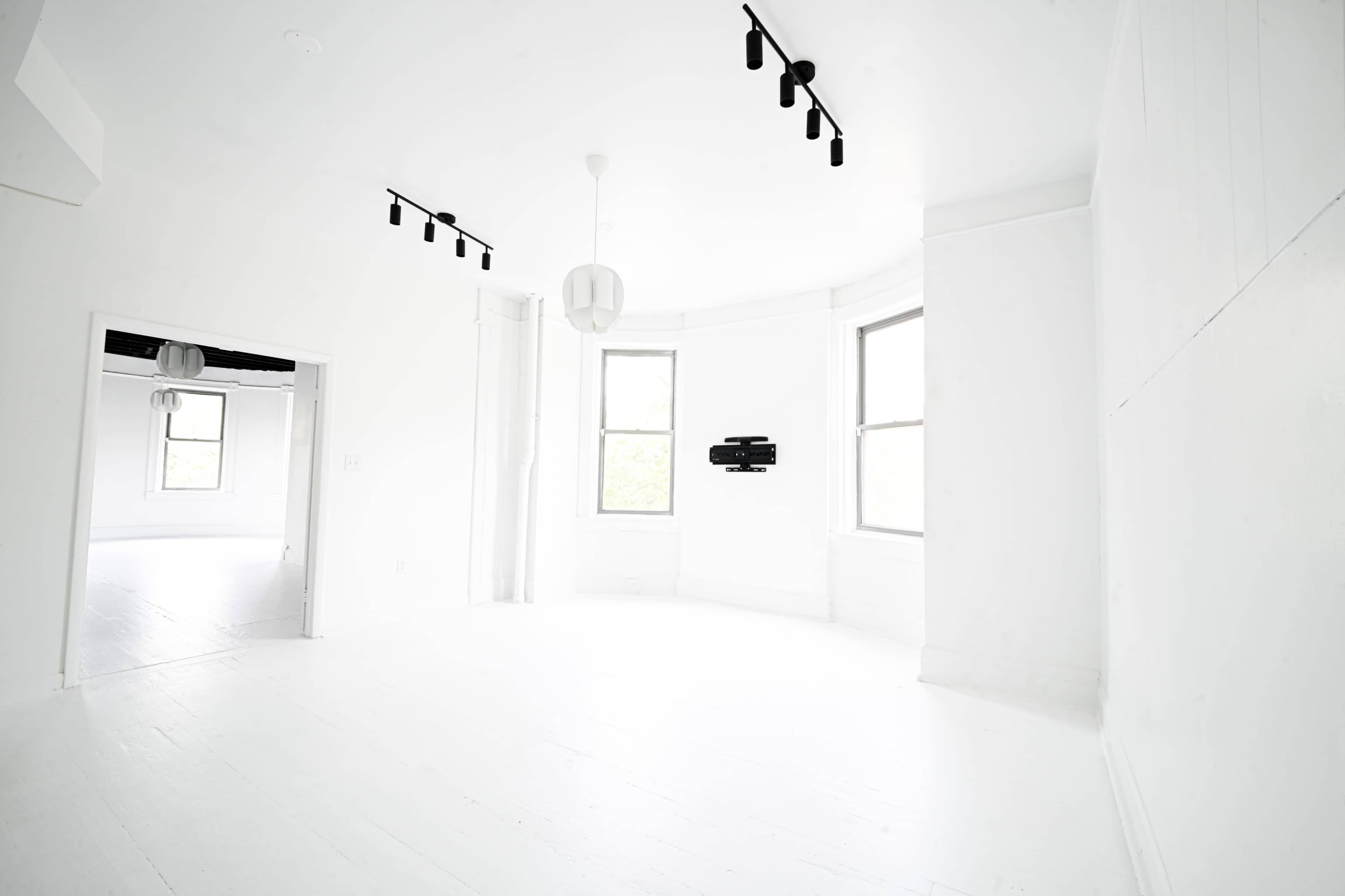 The image shows a spacious, brightly lit room with white walls and flooring, featuring windows and minimalist lighting fixtures.