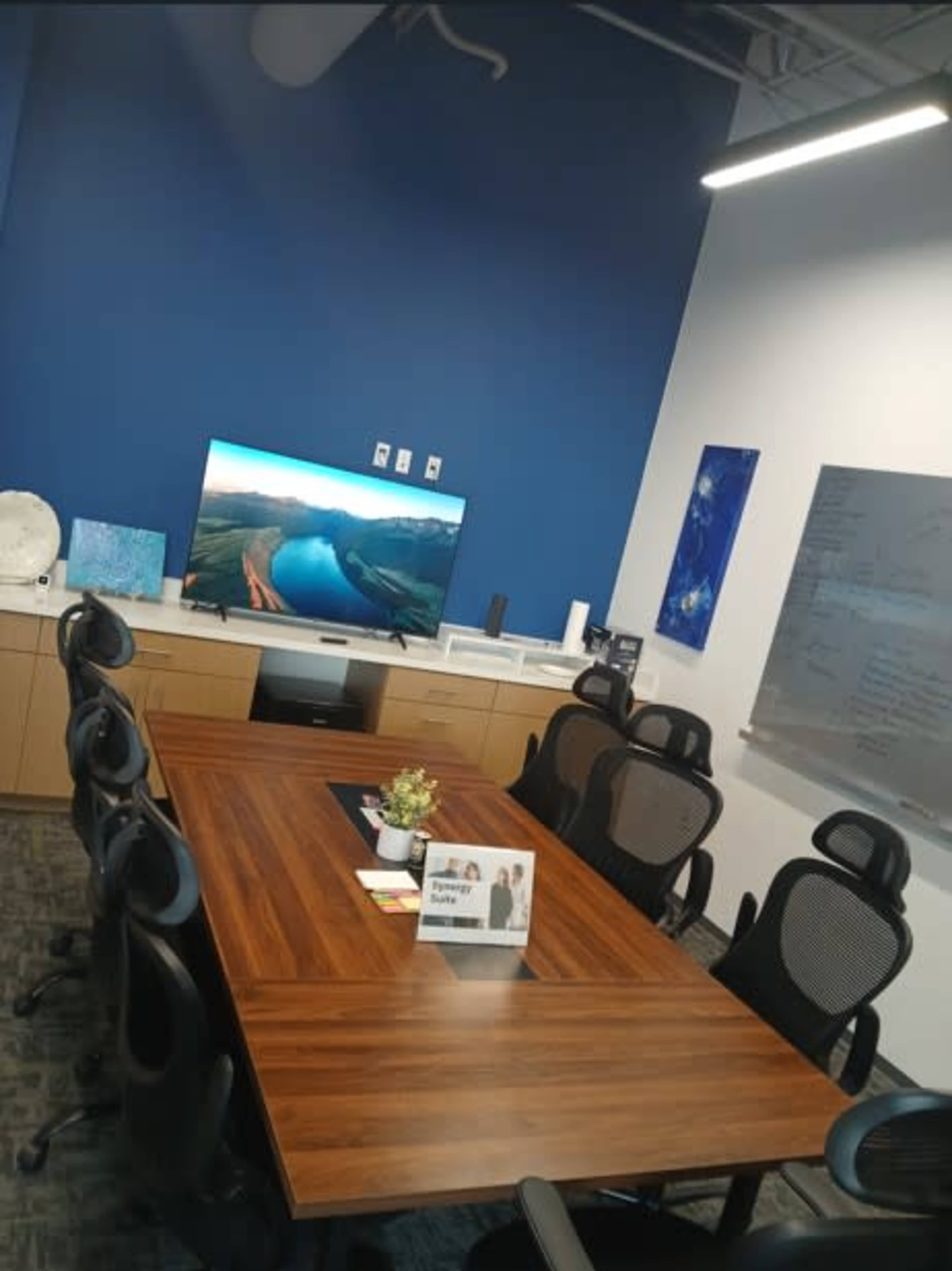 The image shows a conference room with a large wooden table surrounded by ergonomic chairs, a television mounted on the wall, and a whiteboard with notes.