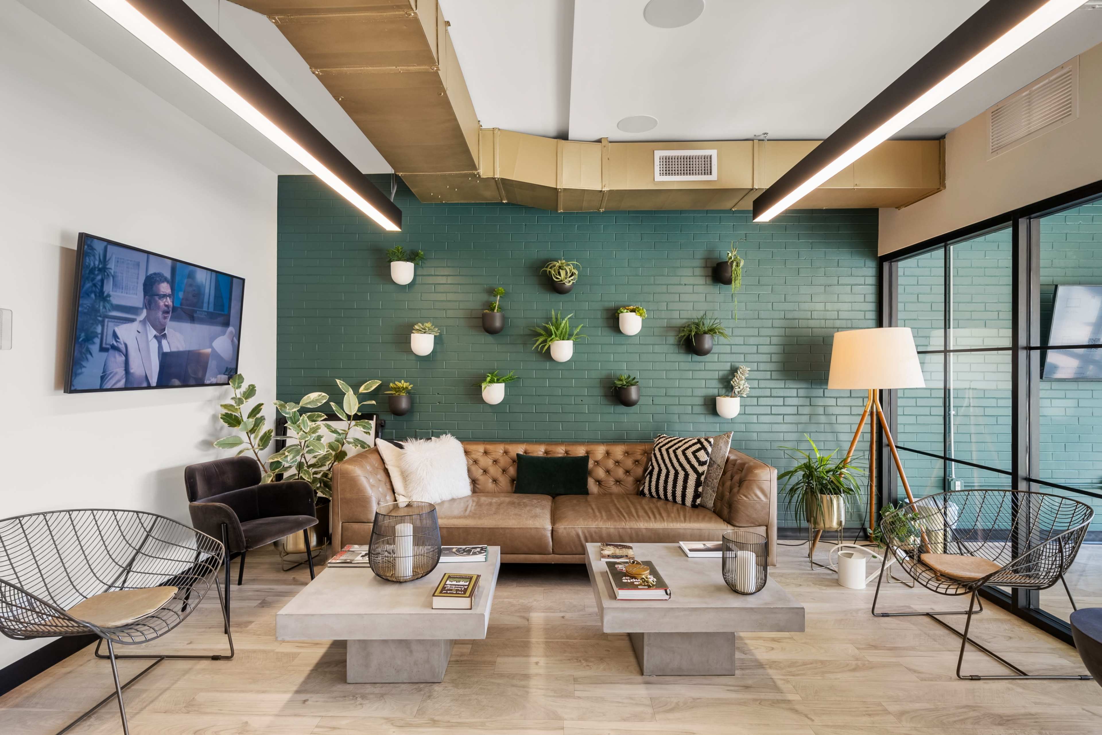 The image shows a modern lounge area with a brown leather sectional couch, concrete coffee tables, and a green wall adorned with potted plants.