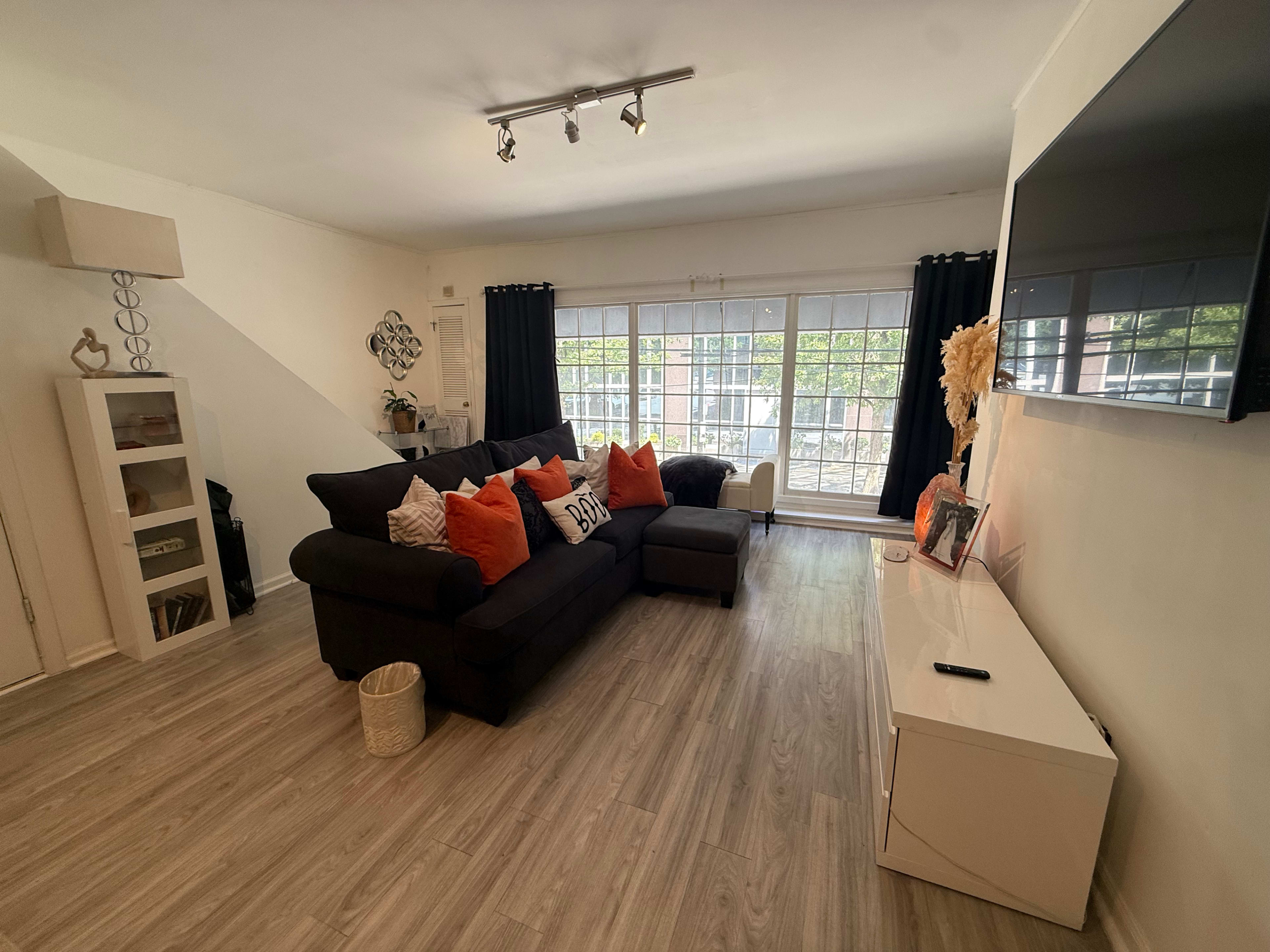 The image shows a modern living room with a black sofa, orange and white decorative pillows, a white media console, and large windows with black curtains, allowing natural light to enter.