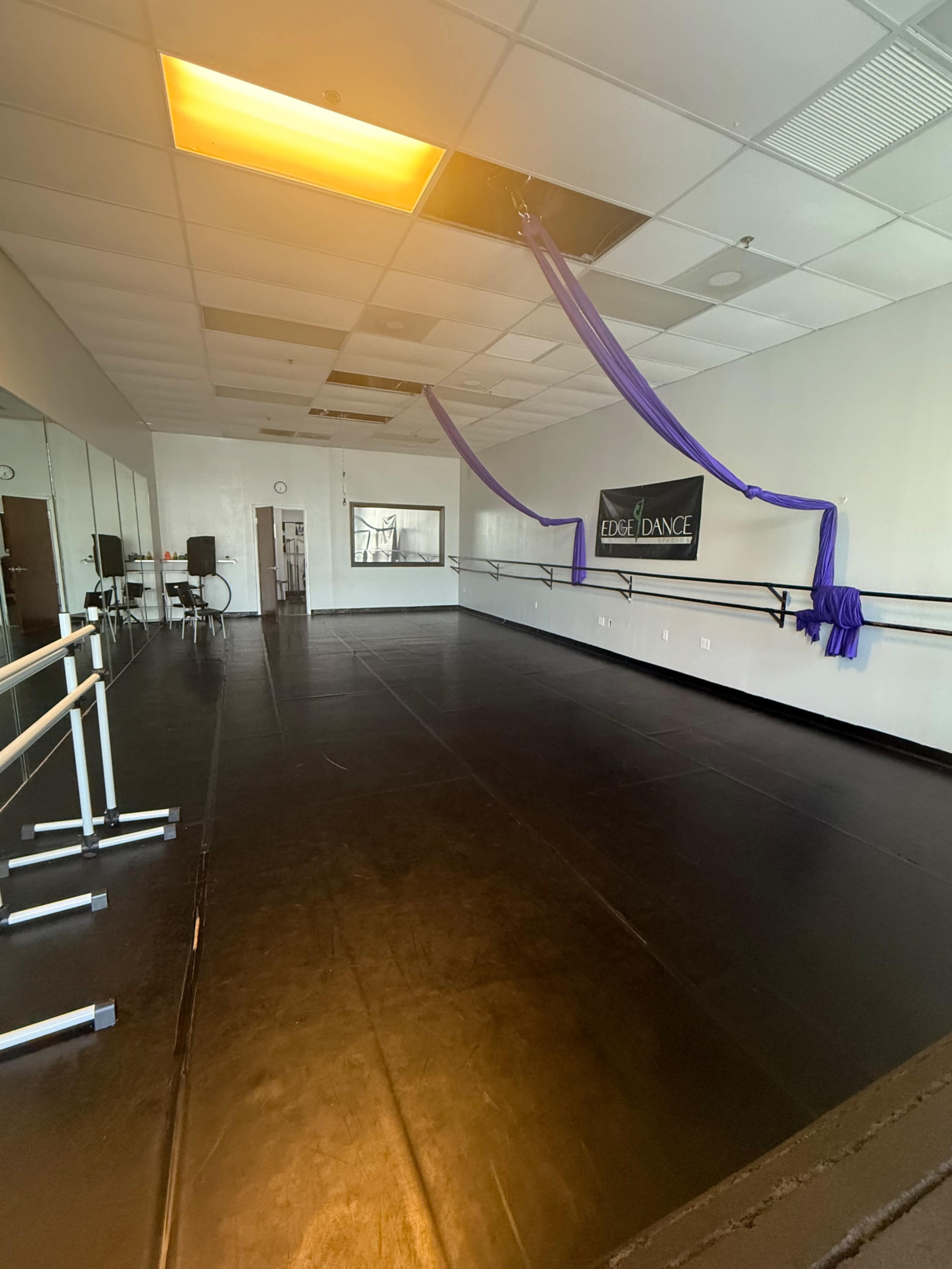 The image shows a dance studio with a black floor, mirrored walls, and a suspended purple fabric for aerial exercises.