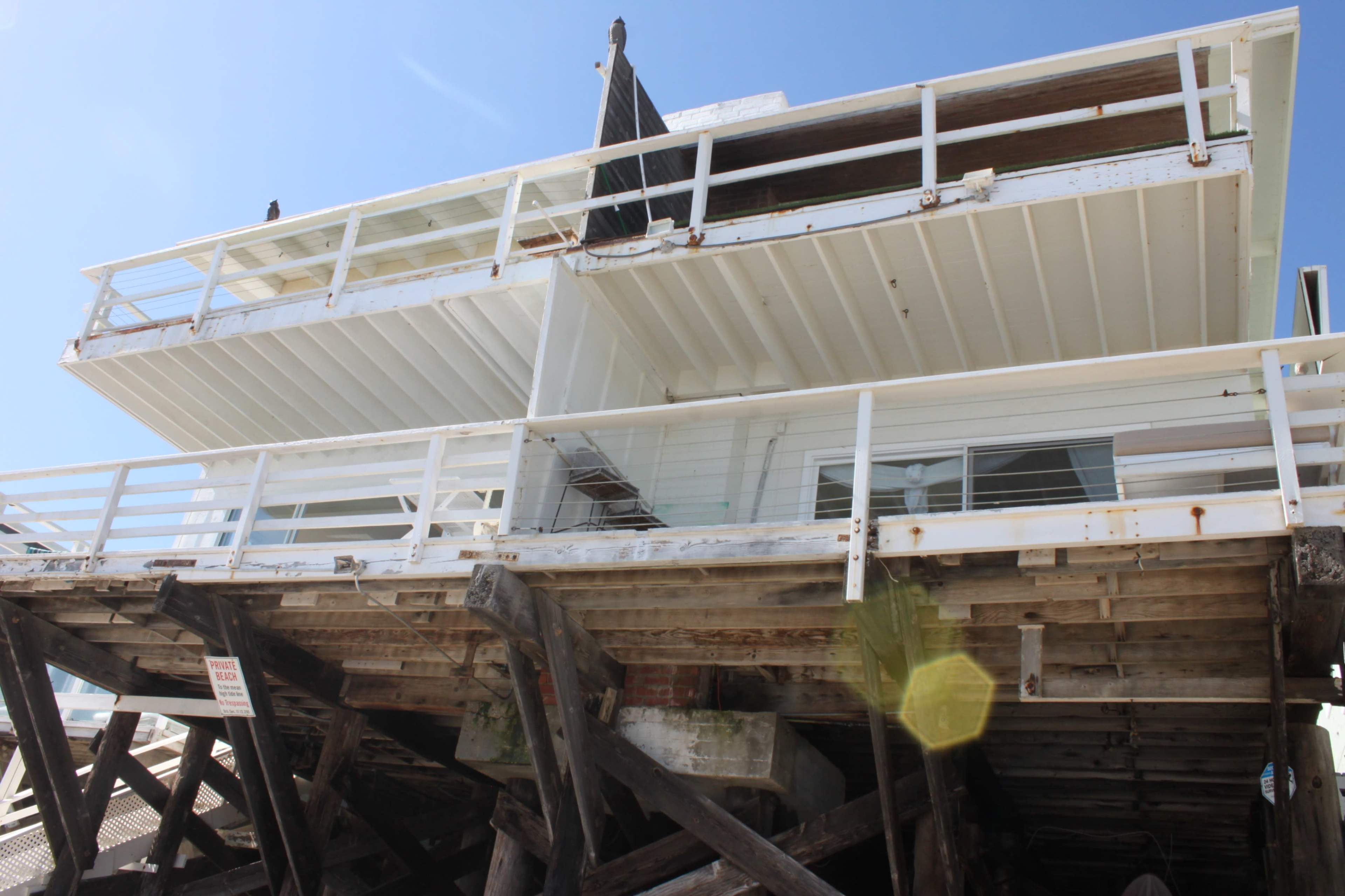 A white beach house is elevated on wooden stilts, with a spacious deck visible against a clear blue sky.