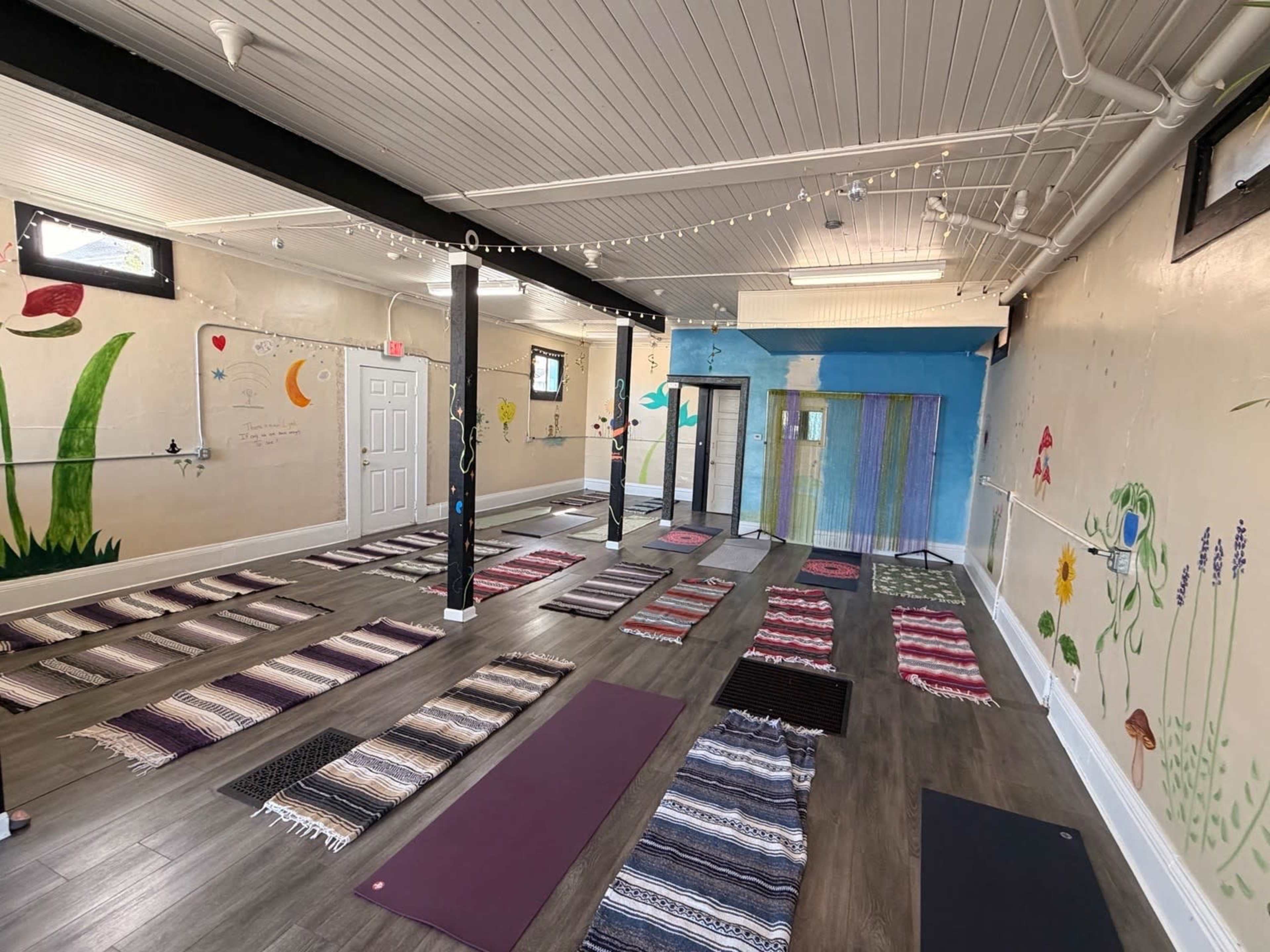 The image shows a yoga studio with several yoga mats arranged on the floor, colorful murals on the walls, and natural lighting coming from windows.